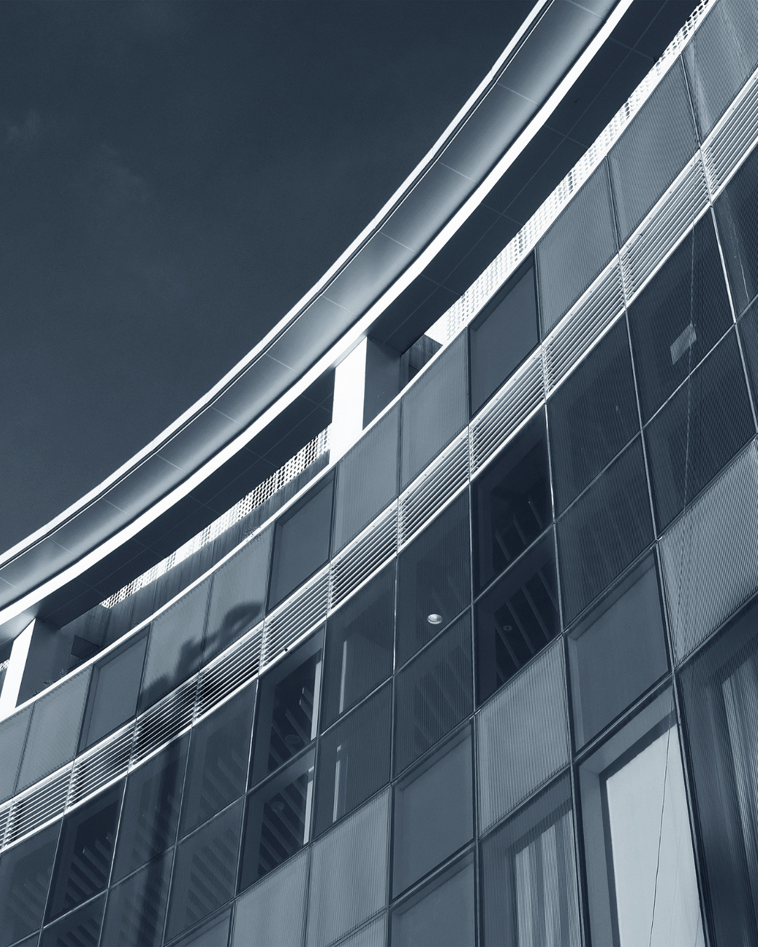 Modern glass building with reflective windows and metal railings, viewed from below against the sky.