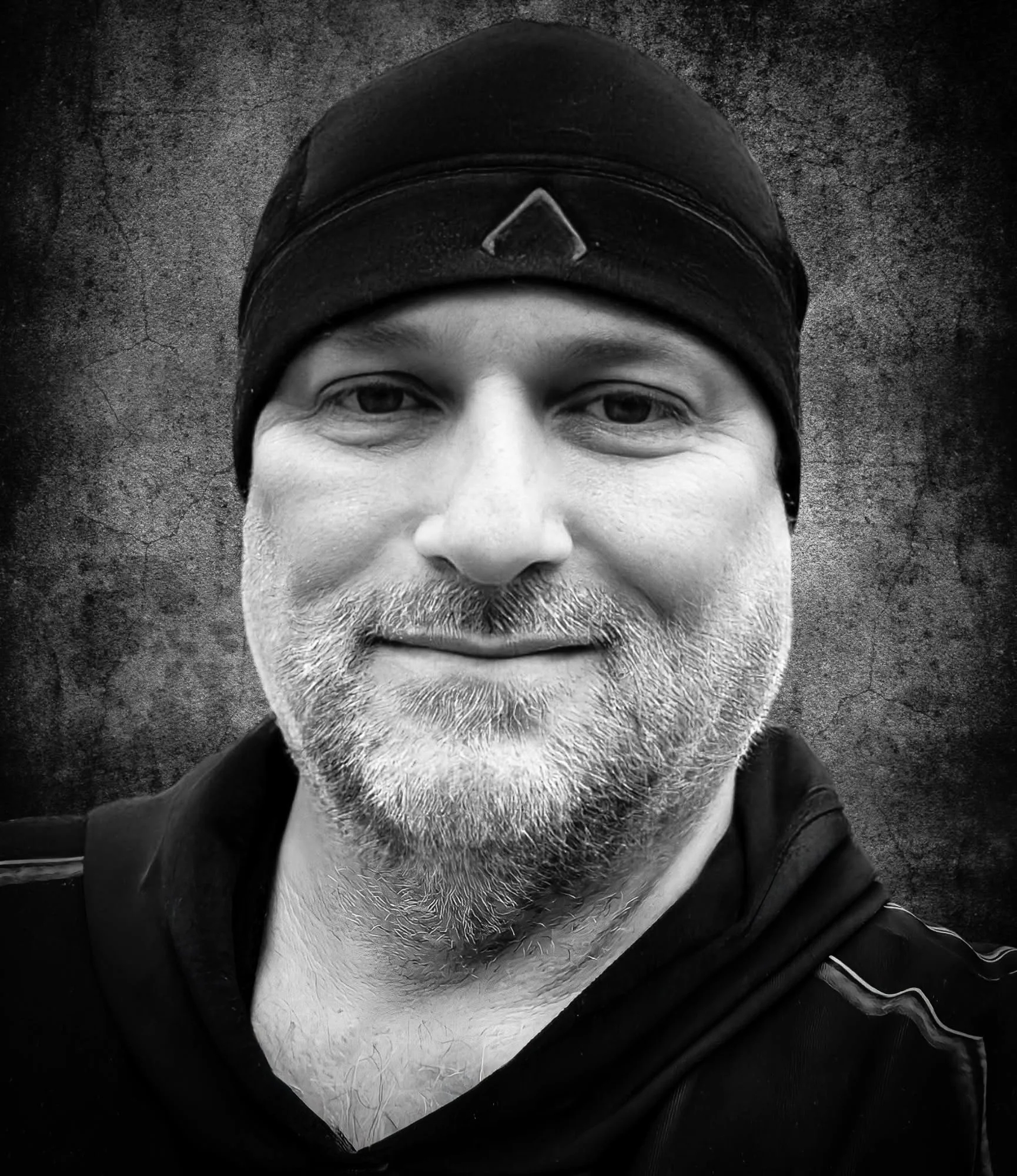 A black and white close-up photo of a man's face wearing a black cap with a triangle emblem, against a textured dark background.