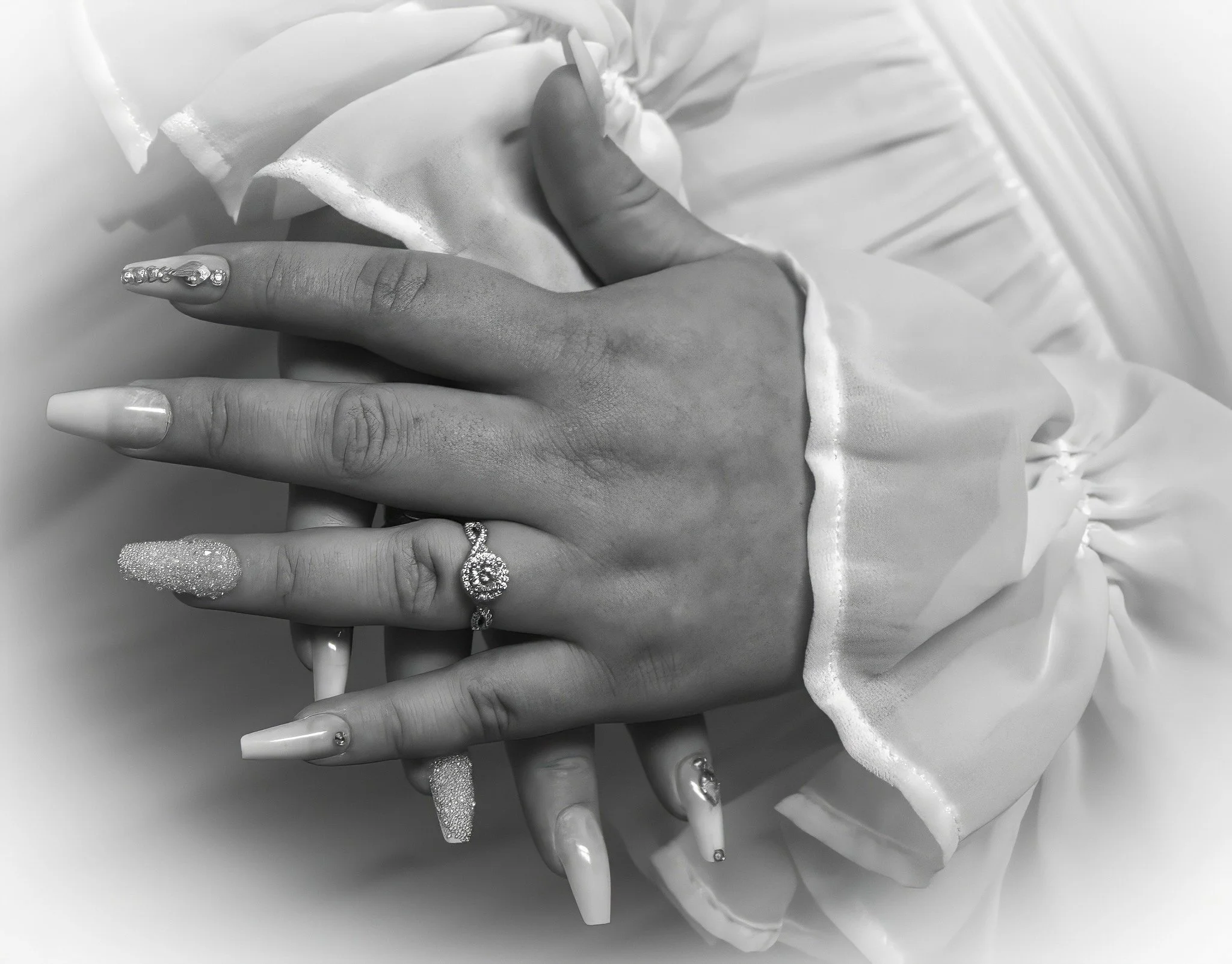 Close-up of hands with diamond rings and decorated nails, resting on fabric with ruffled edges.