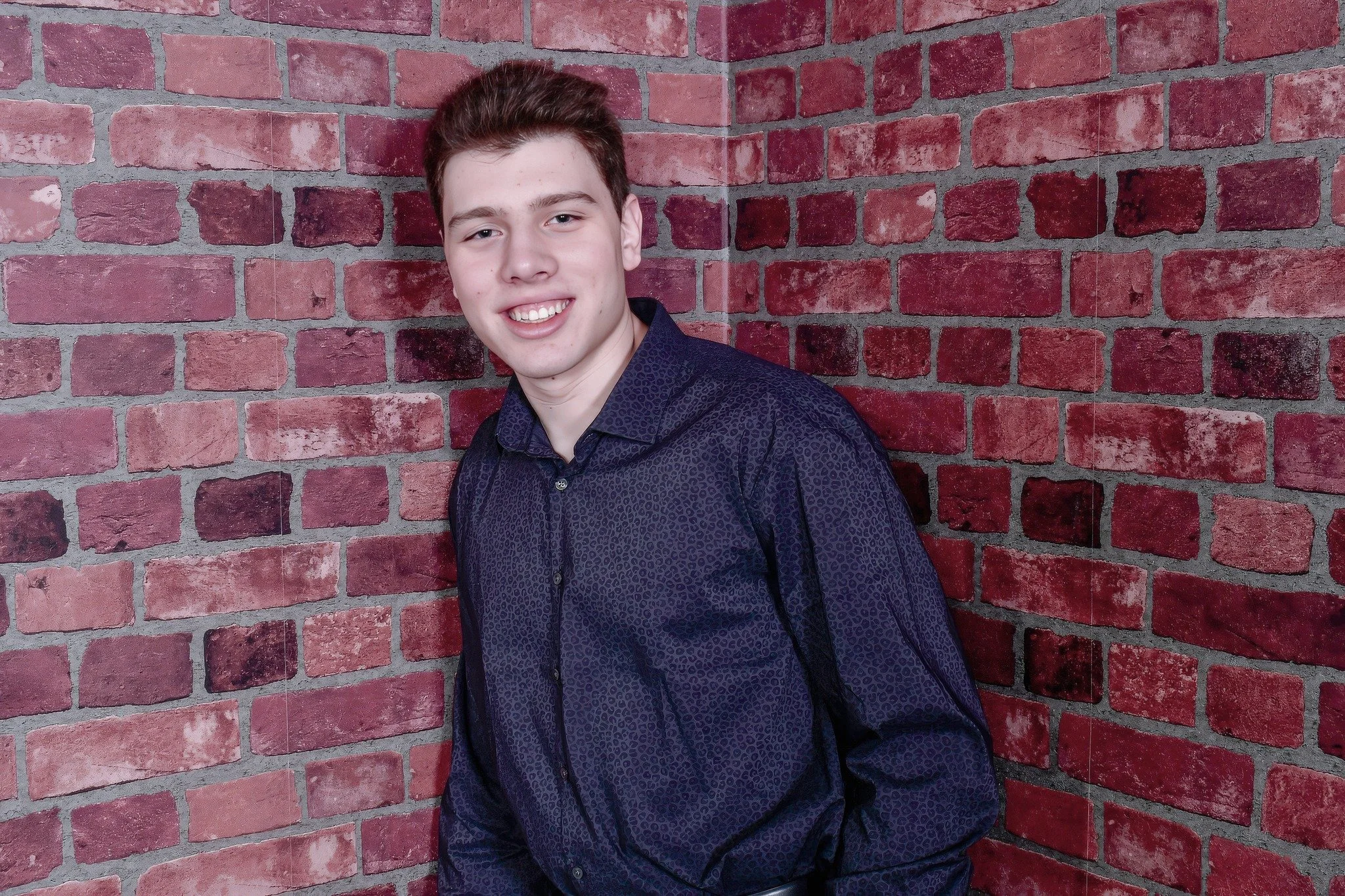 A young man with short brown hair, wearing a dark patterned shirt, smiling while leaning against a red brick wall.
