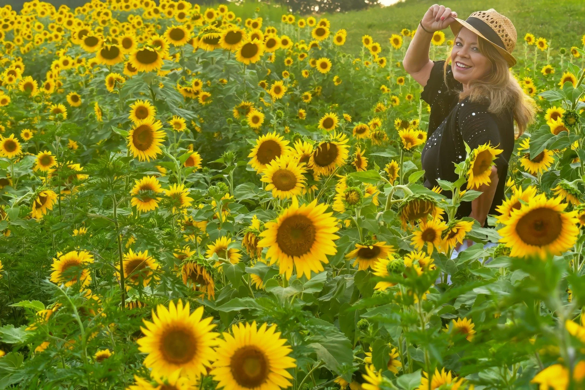 A smiling woman wearing a straw hat standing in a sunflower field.