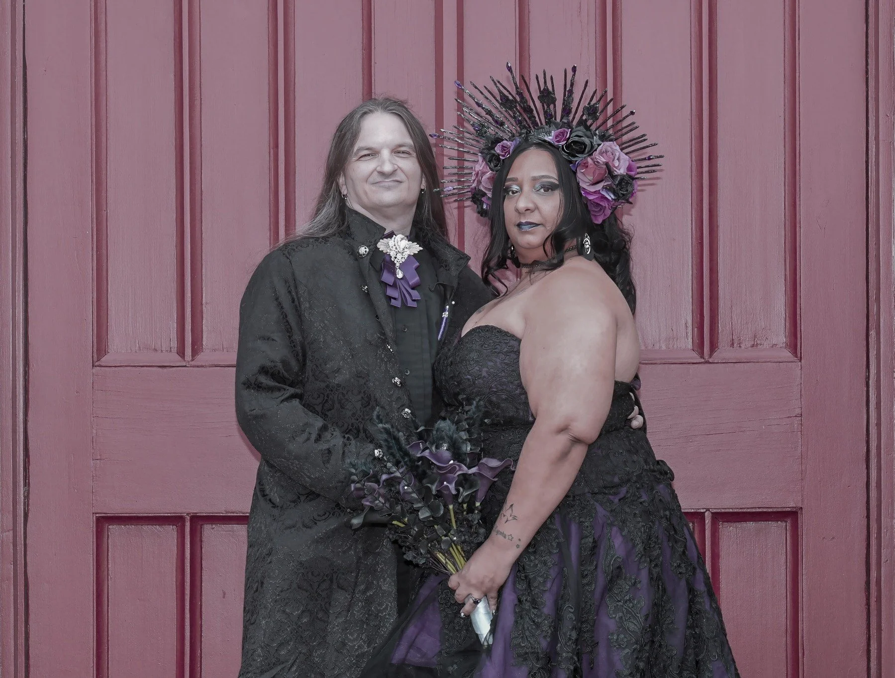 A man and woman dressed in black standing in front of a pink door. The woman wears a gothic style dress with a large floral and spiked crown, and holds a bouquet of dark purple flowers. The man wears a black jacket with lace details and decorative pins, with long hair and an earring.