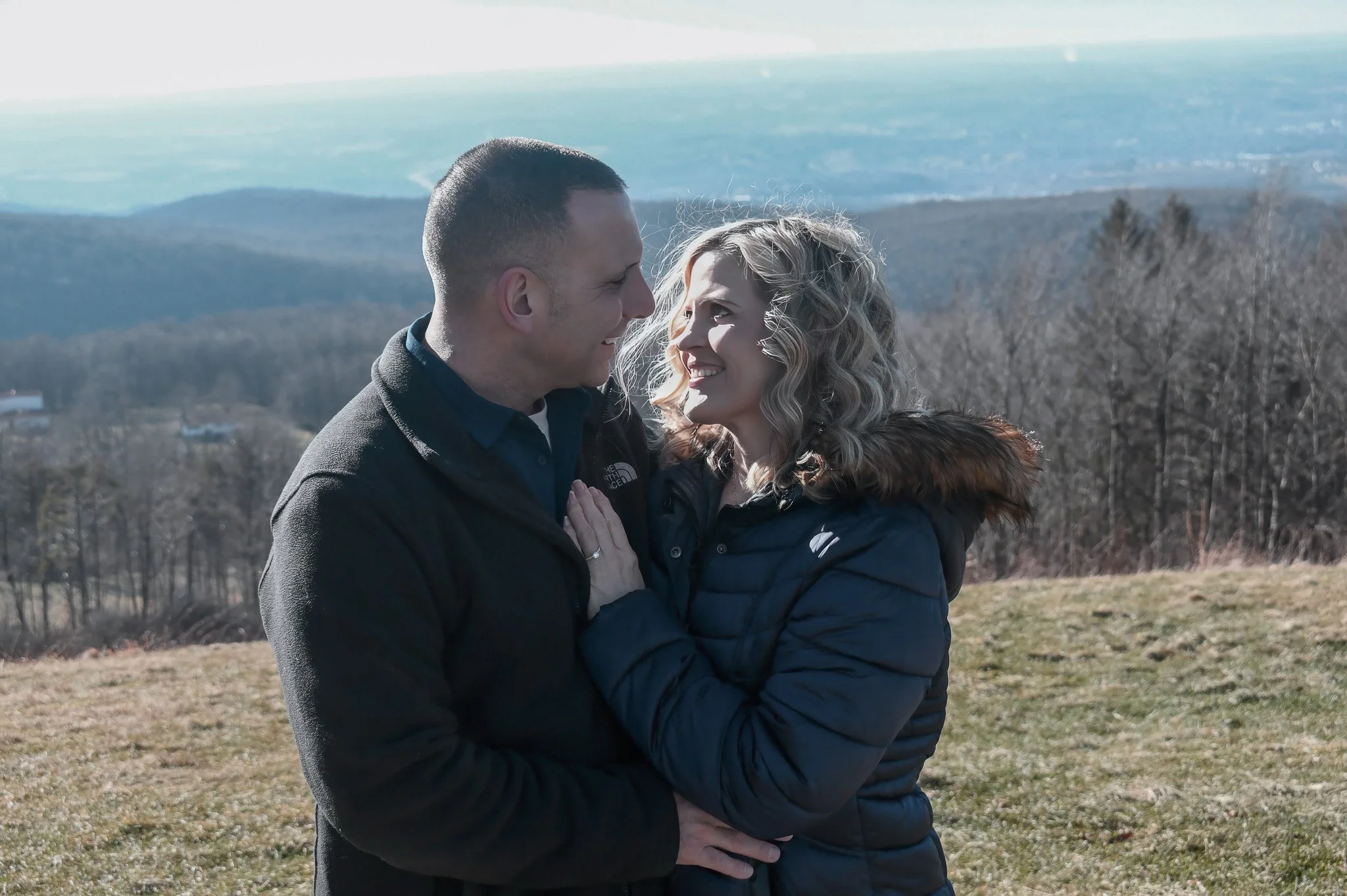 A couple standing outdoors facing each other, smiling, with their foreheads touching, in a scenic landscape with distant mountains and leafless trees.