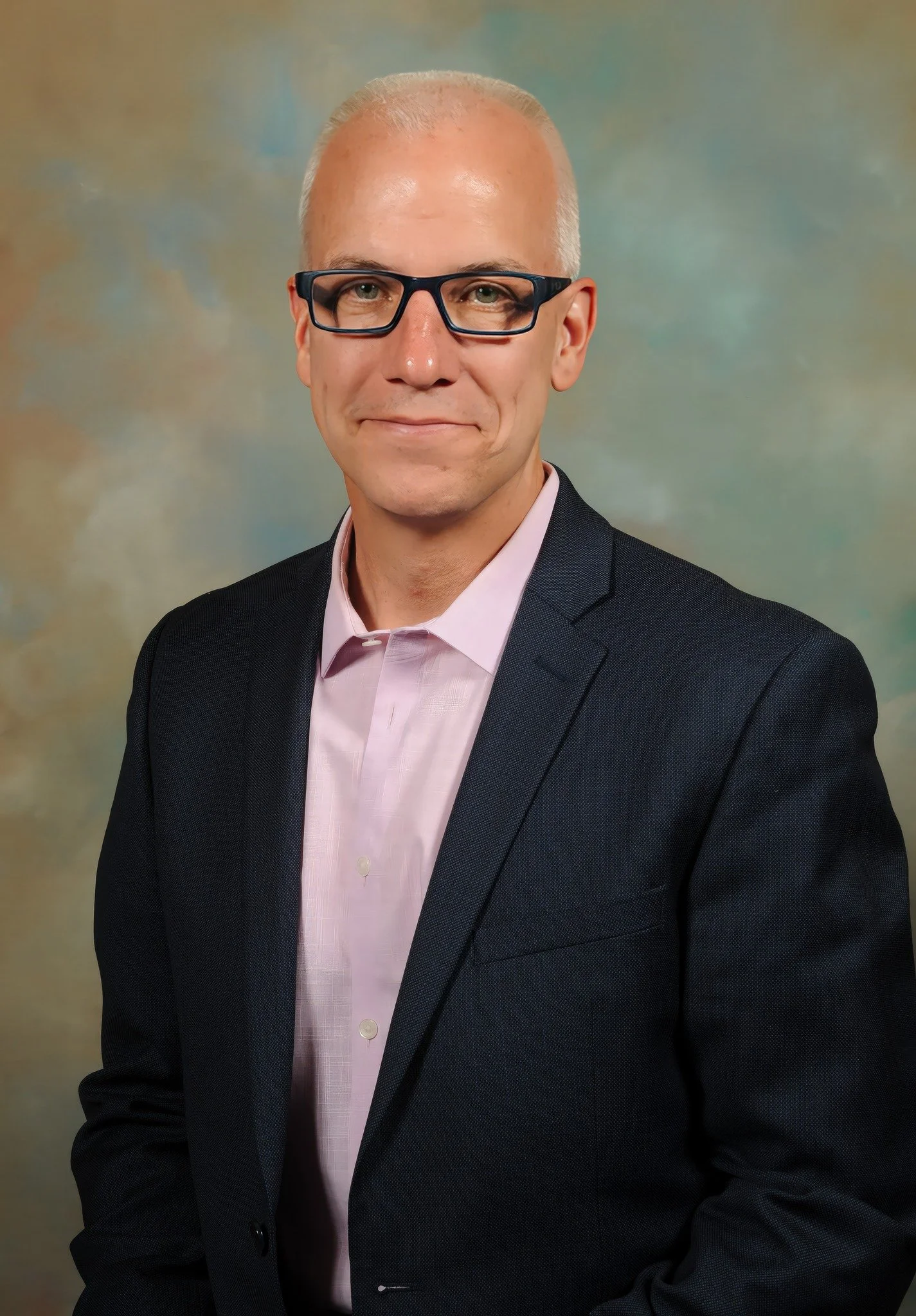 A middle-aged man with short, gray hair and glasses, wearing a black suit jacket and a light pink dress shirt, smiling in a professional headshot against a blurred, multicolored background.