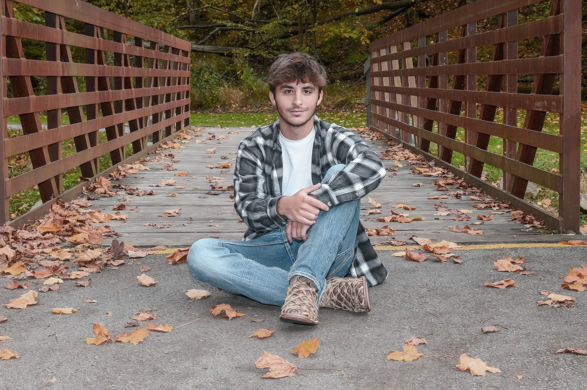 A young man sitting on a wooden bridge covered with fallen autumn leaves, wearing a checkered shirt and jeans, with a forested background.