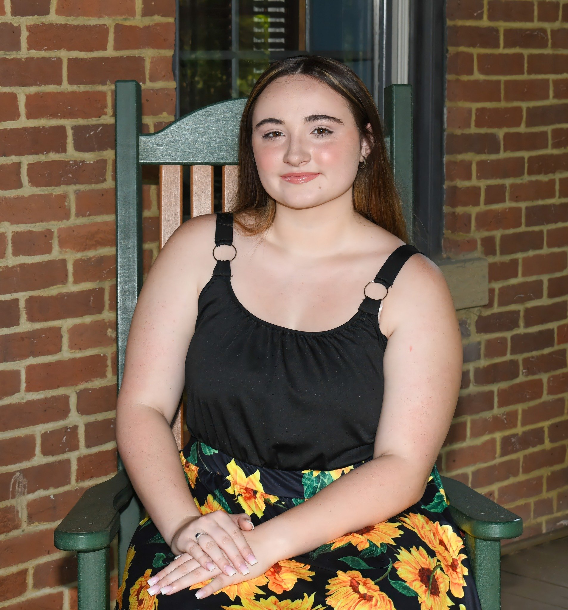 A young woman with light skin, dark brown hair, and brown eyes, smiling while sitting on a green wooden chair outside in front of a brick wall, wearing a black dress with sunflower and tropical floral prints.