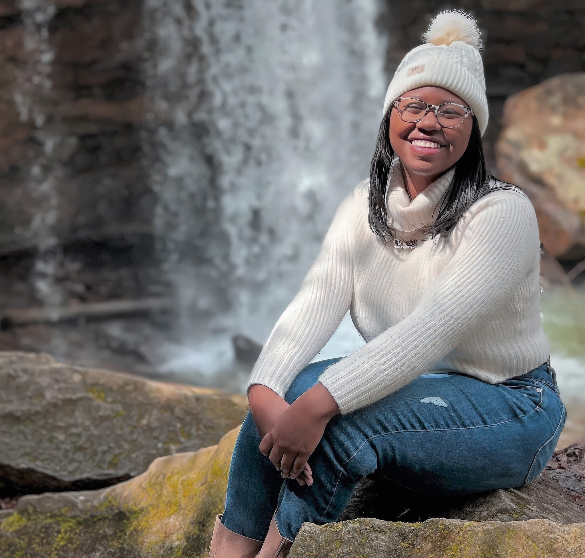 A smiling woman wearing glasses, a white beanie with a pom-pom, and a light-colored sweater sitting outdoors near a waterfall.