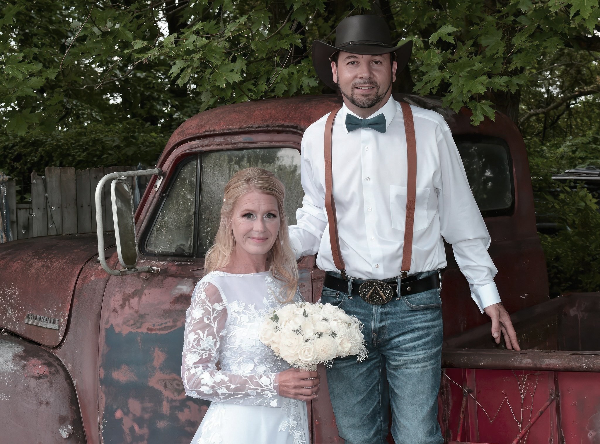A man and woman stand in front of an old red truck with a leafy green background. The woman is dressed in a white wedding dress holding a bouquet, and the man is wearing a white shirt, bow tie, suspenders, and a cowboy hat.
