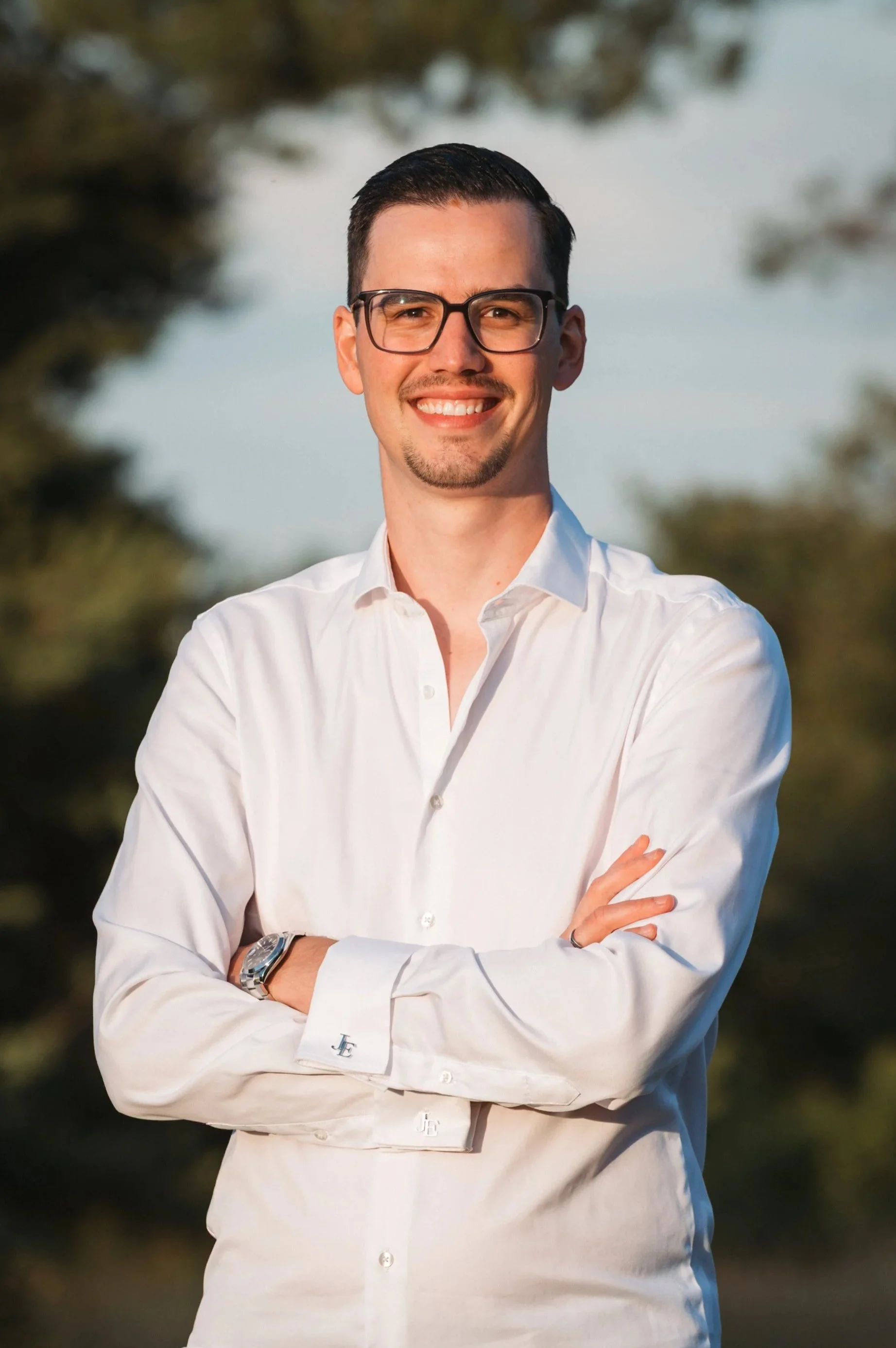 Ein Mann mit dunklem Haar, Brille und Bart trägt ein weißes Hemd, steht vor uns und lächelt. Er hat seine Arme verschränkt, trägt eine Armbanduhr und befindet sich im Freien mit unscharfem Naturhintergrund.