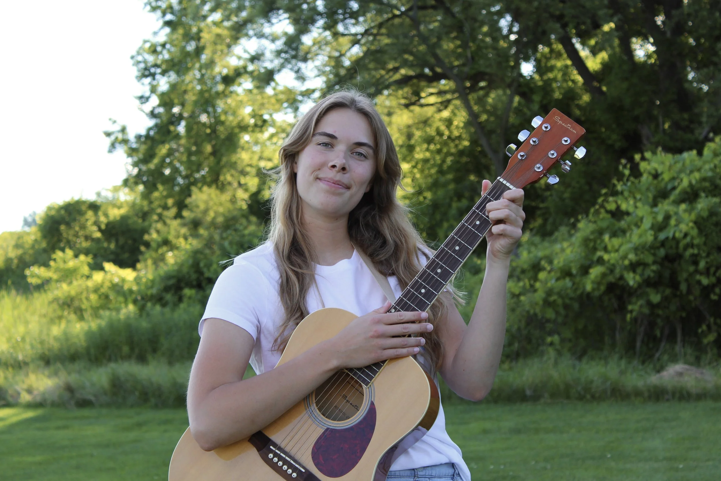 A young woman with light brown hair and a white shirt holding an acoustic guitar outdoors surrounded by green trees and bushes.