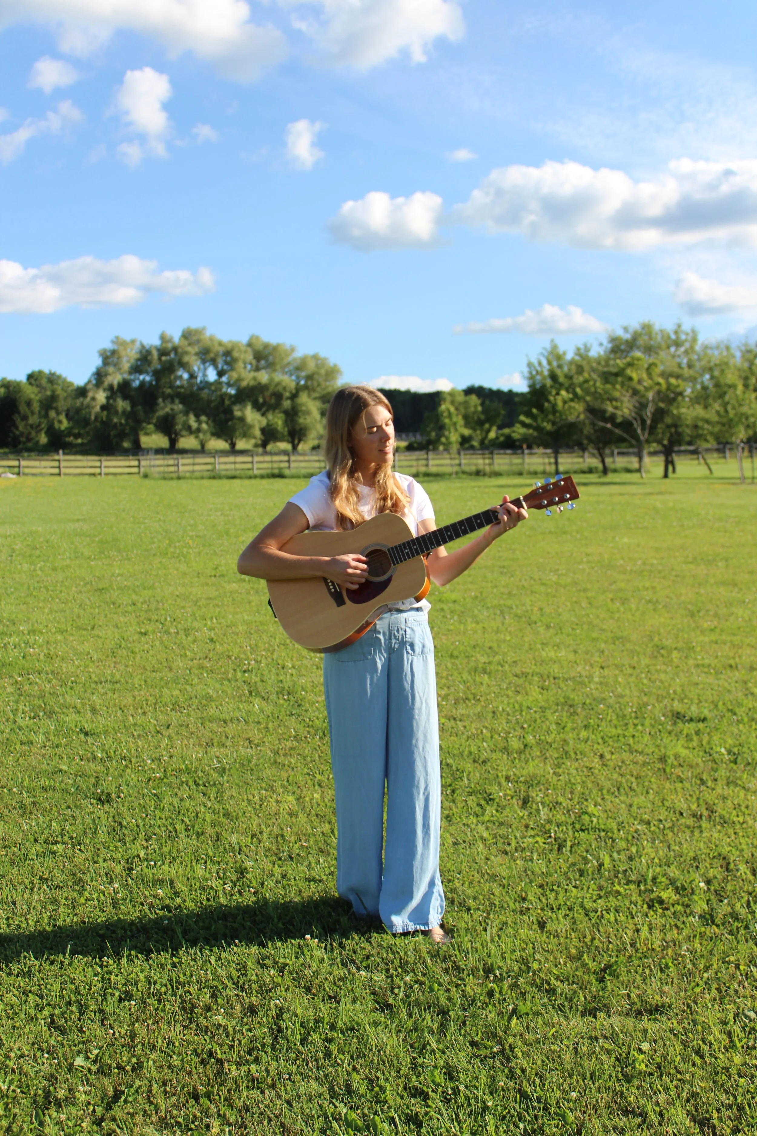 A young woman with long blonde hair, wearing a white T-shirt and light blue wide-leg pants, standing in a grassy field and playing an acoustic guitar under a blue sky with scattered clouds.