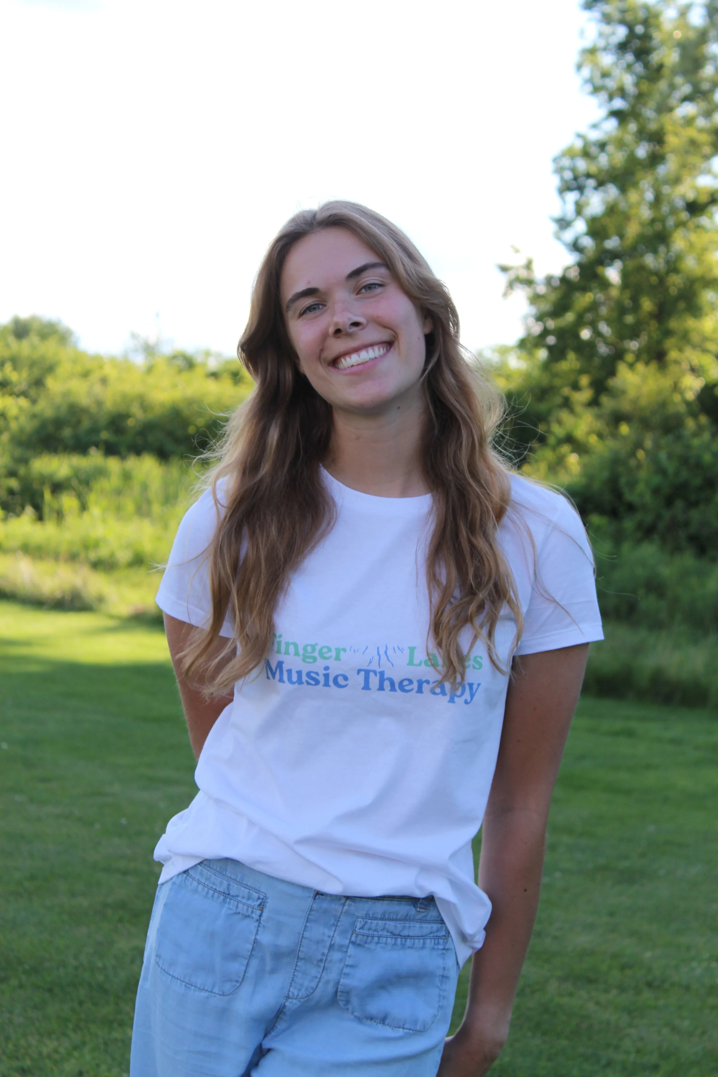 A young woman with long wavy brown hair smiling outdoors in a park with green trees and grass in the background. She is wearing a white t-shirt with text related to music therapy and light blue pants.