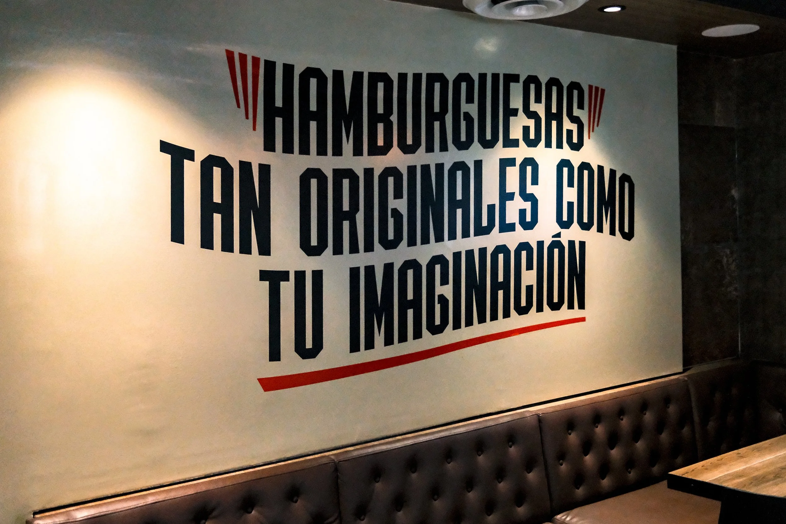 A wall painted with the quote "Hamburguesas Tan Originales Como Tu Imaginación" above a leather bench, by OLA Creative Studio.