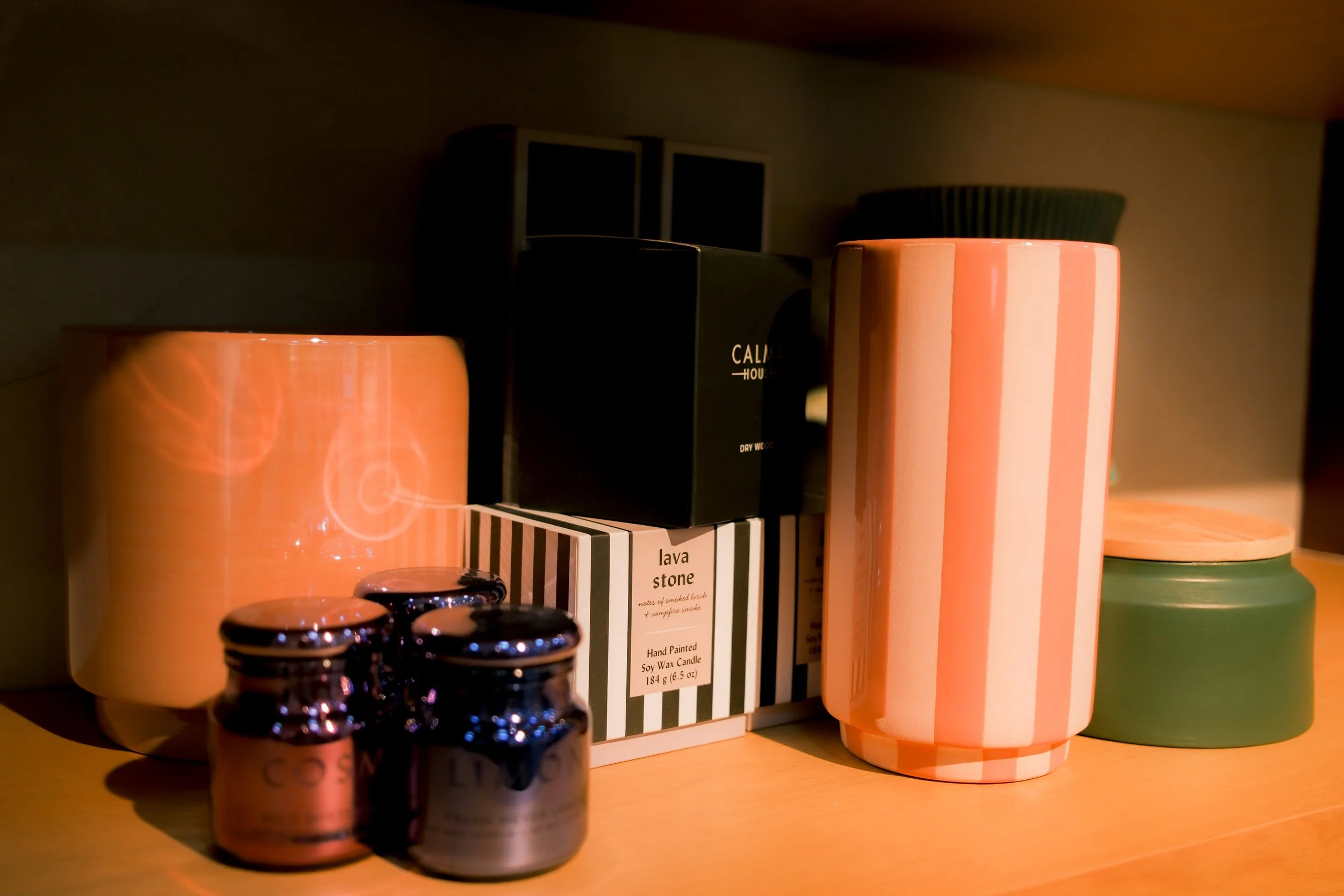 A close-up of candles and black bottles styled on a shelf, including a pink striped candle and a box of lava stone, by OLA Creative Studio