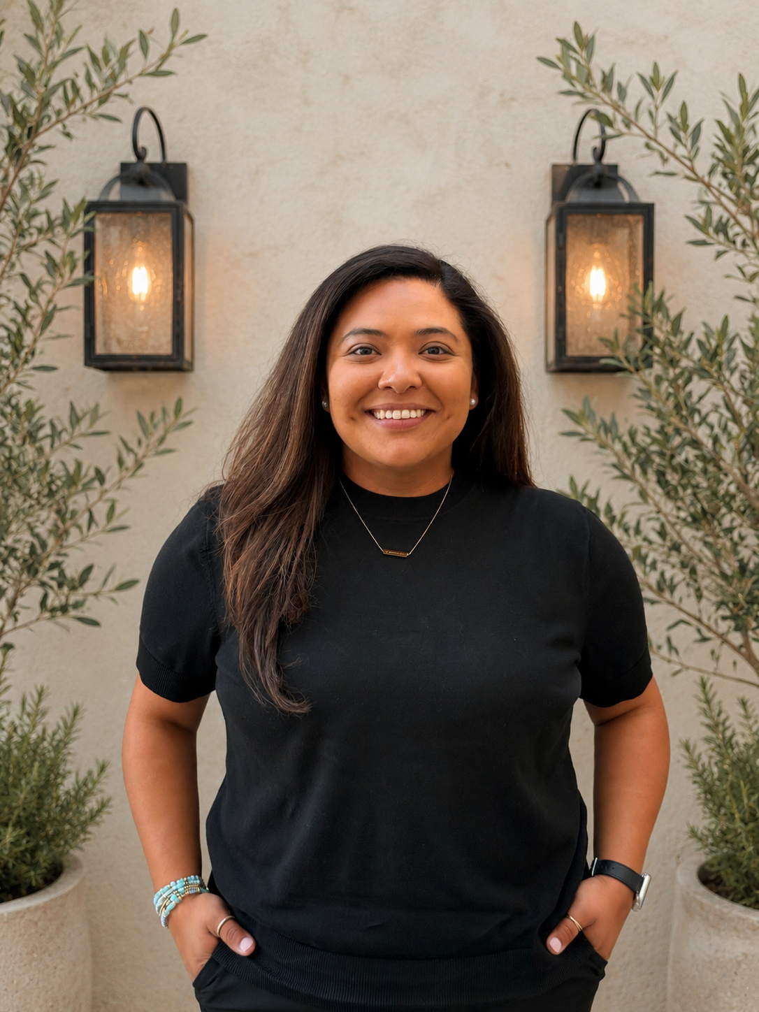 A woman standing outdoors in front of a beige wall with two black outdoor wall lanterns. She has long dark hair, is smiling, and is wearing a black t-shirt, pearl earrings, a necklace, a watch, and bracelets. She has her hands in her pockets.