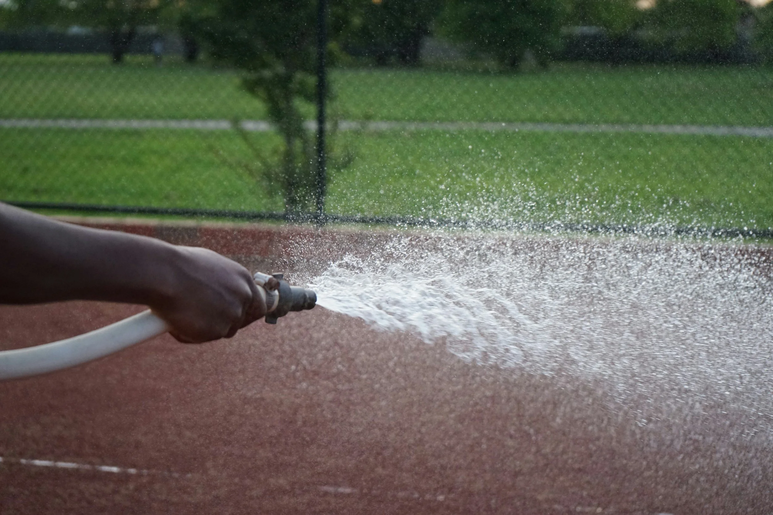 Close-up of a person's hand holding a garden hose and watering a reddish-brown outdoor surface, with a grassy area and trees in the background.