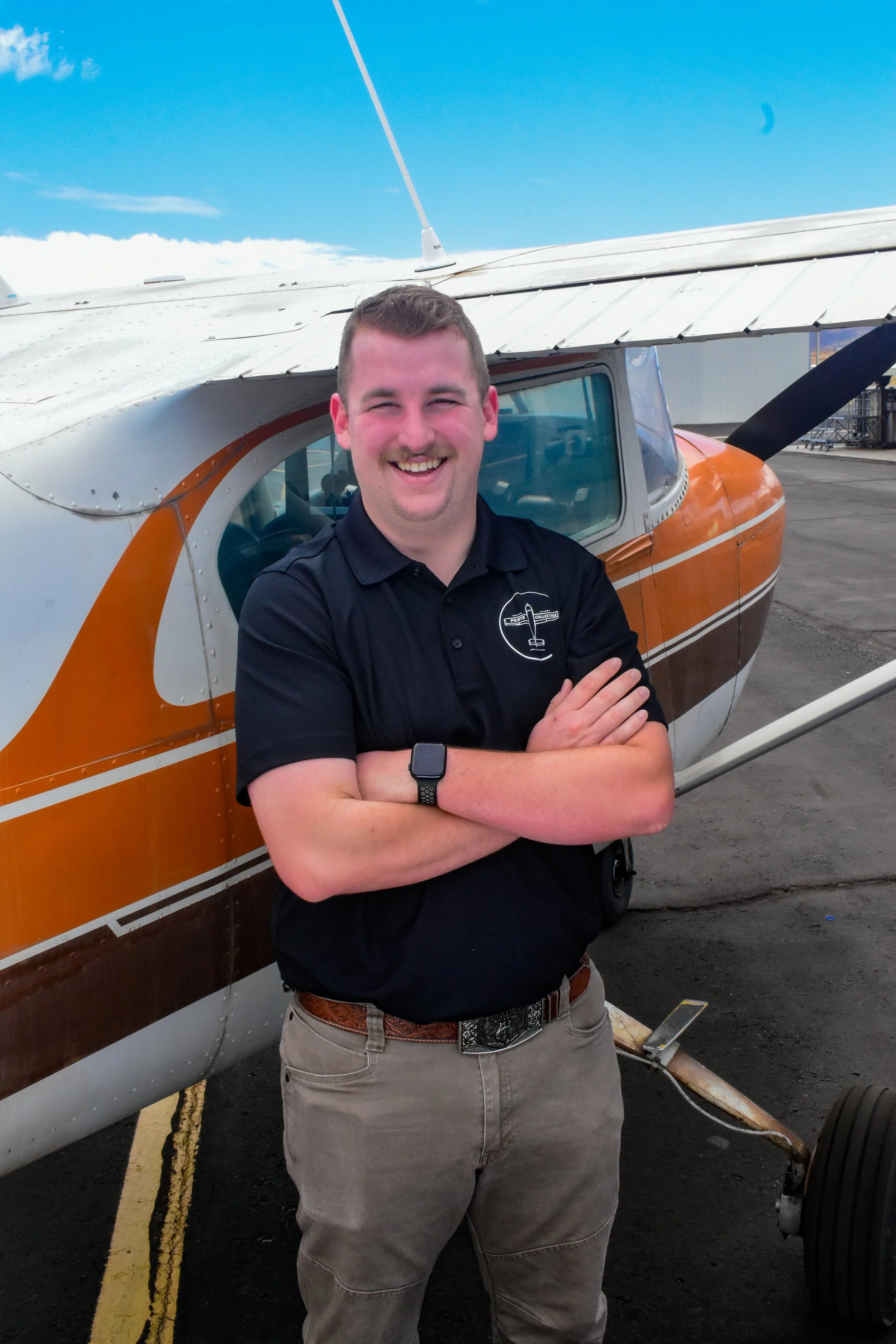 A smiling man with crossed arms standing in front of small aircraft with orange and white paint on the tarmac.