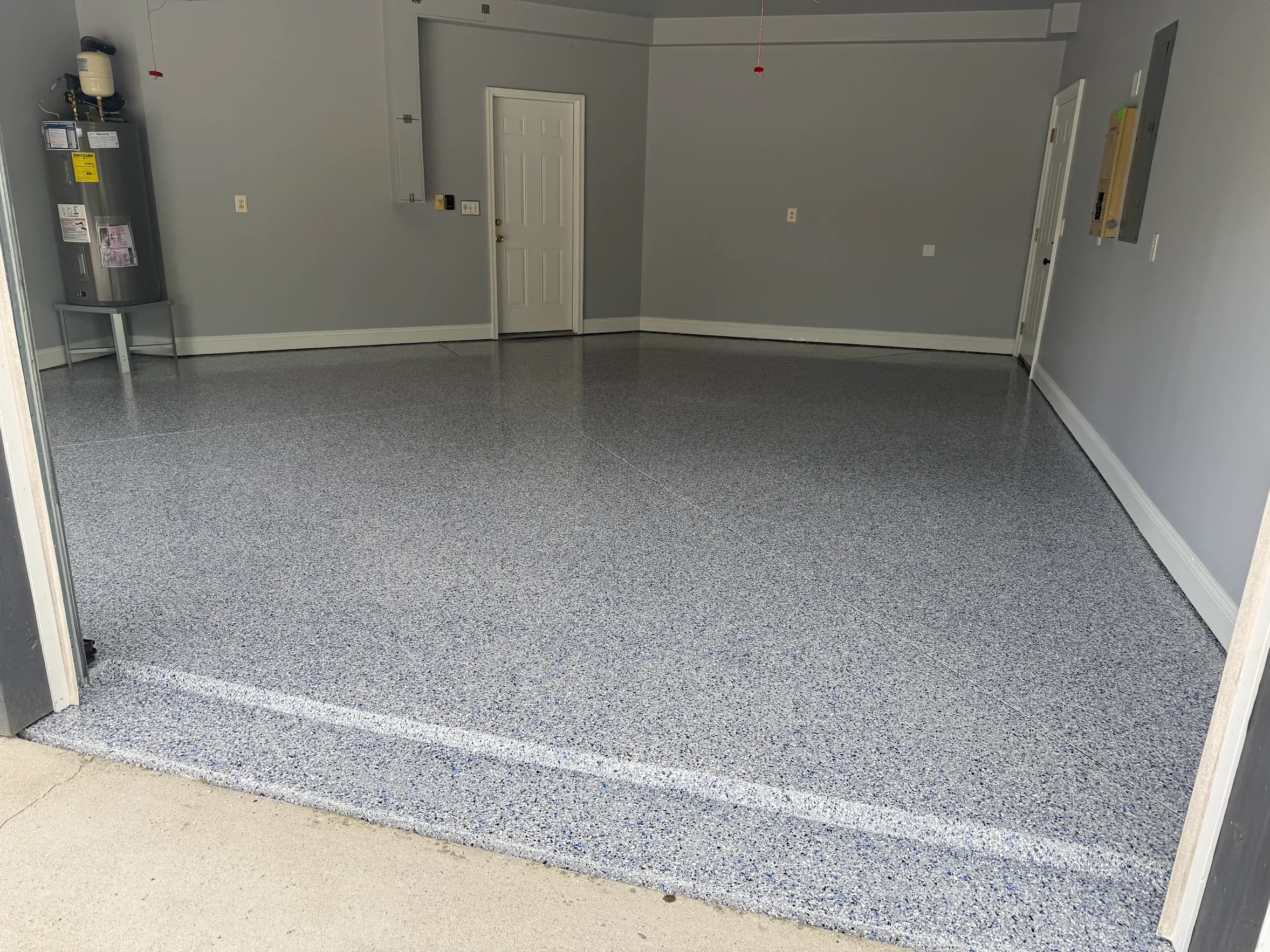 Empty garage with grey speckled epoxy floor, grey walls, a white door, and utility panels on the wall.