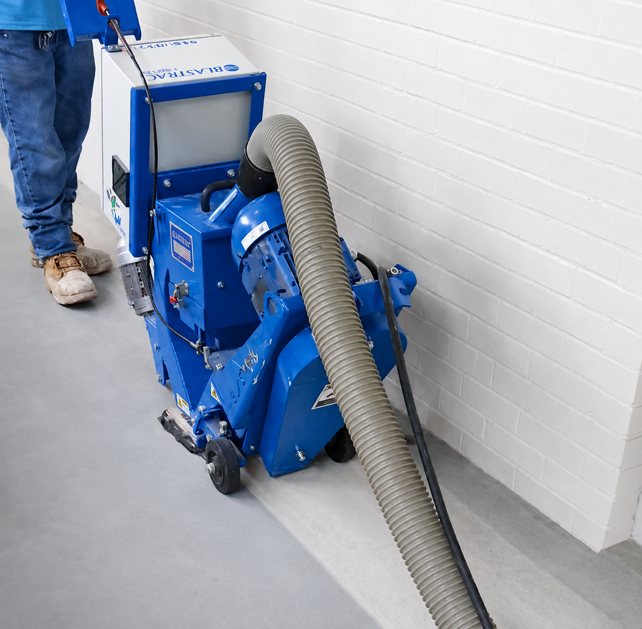 A blue carpet cleaning machine with a large vacuum hose attached, placed against a white brick wall on a gray floor, with a person wearing jeans and boots nearby.