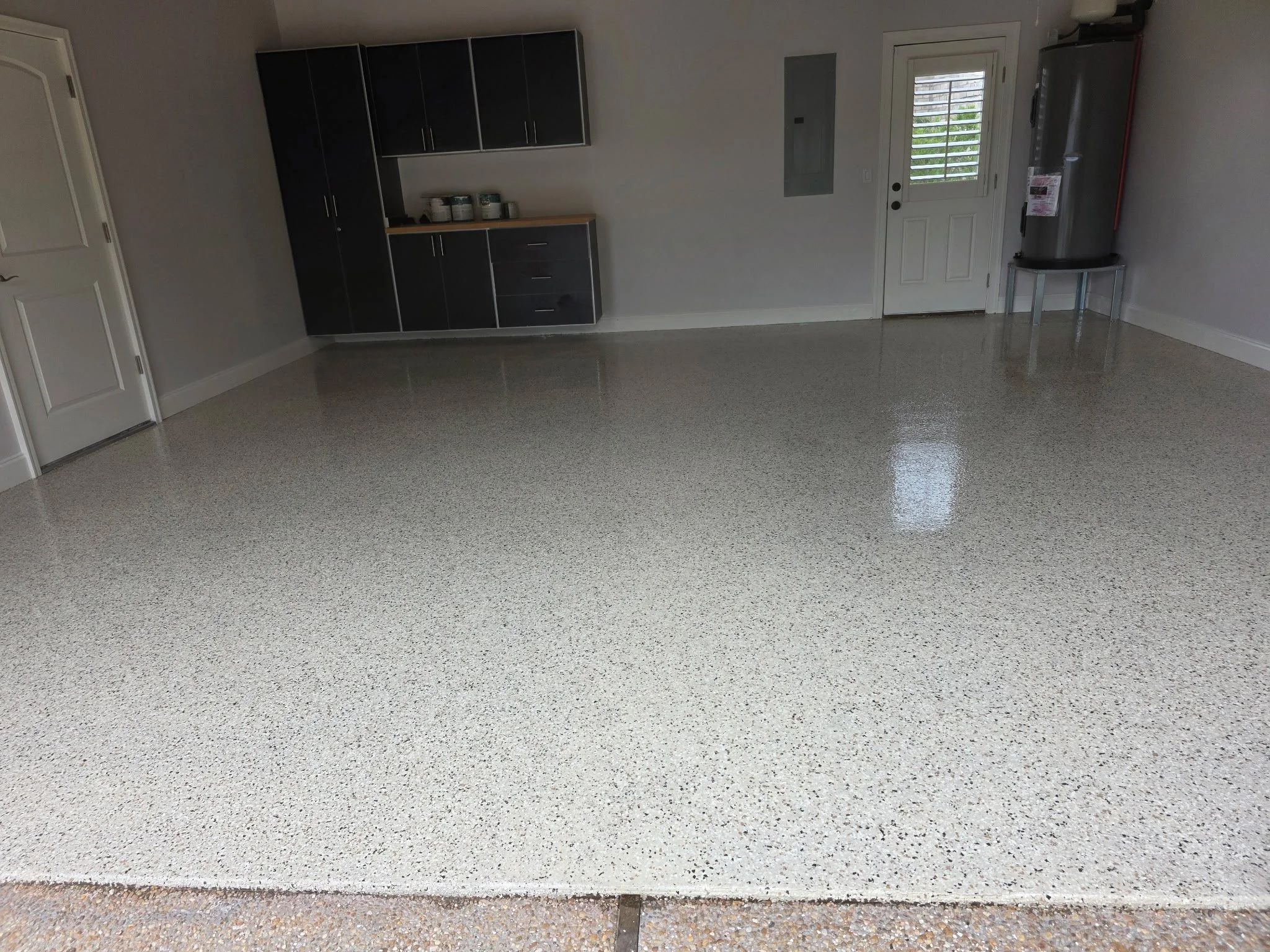 Empty garage with a polished concrete floor, black cabinets on the left wall, a water heater on the right, and a door with a window and shutters.