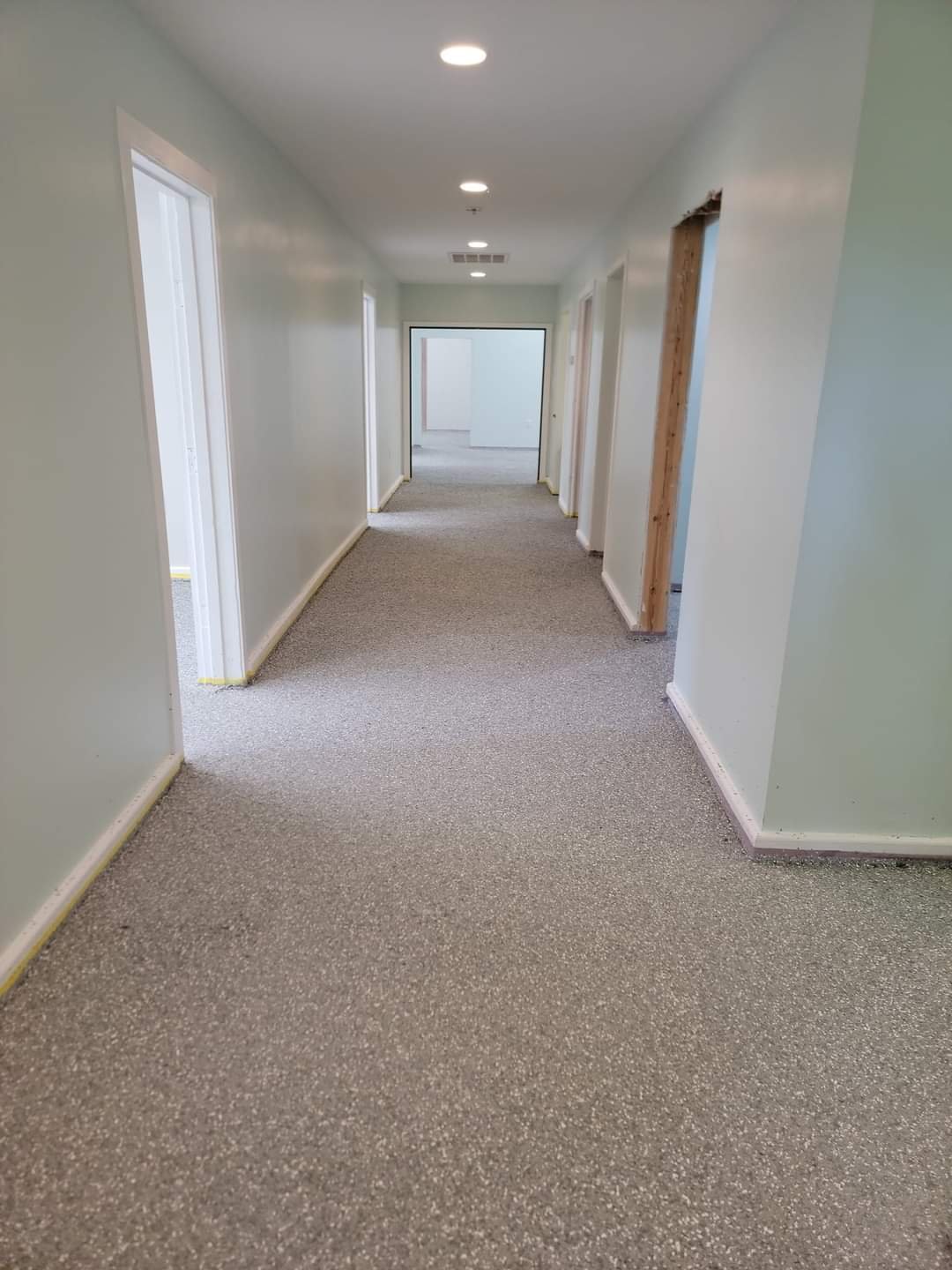 A hallway with painted light green walls, carpeted flooring, and ceiling lights. Several doorways are visible along the hallway, some framed and unfinished.