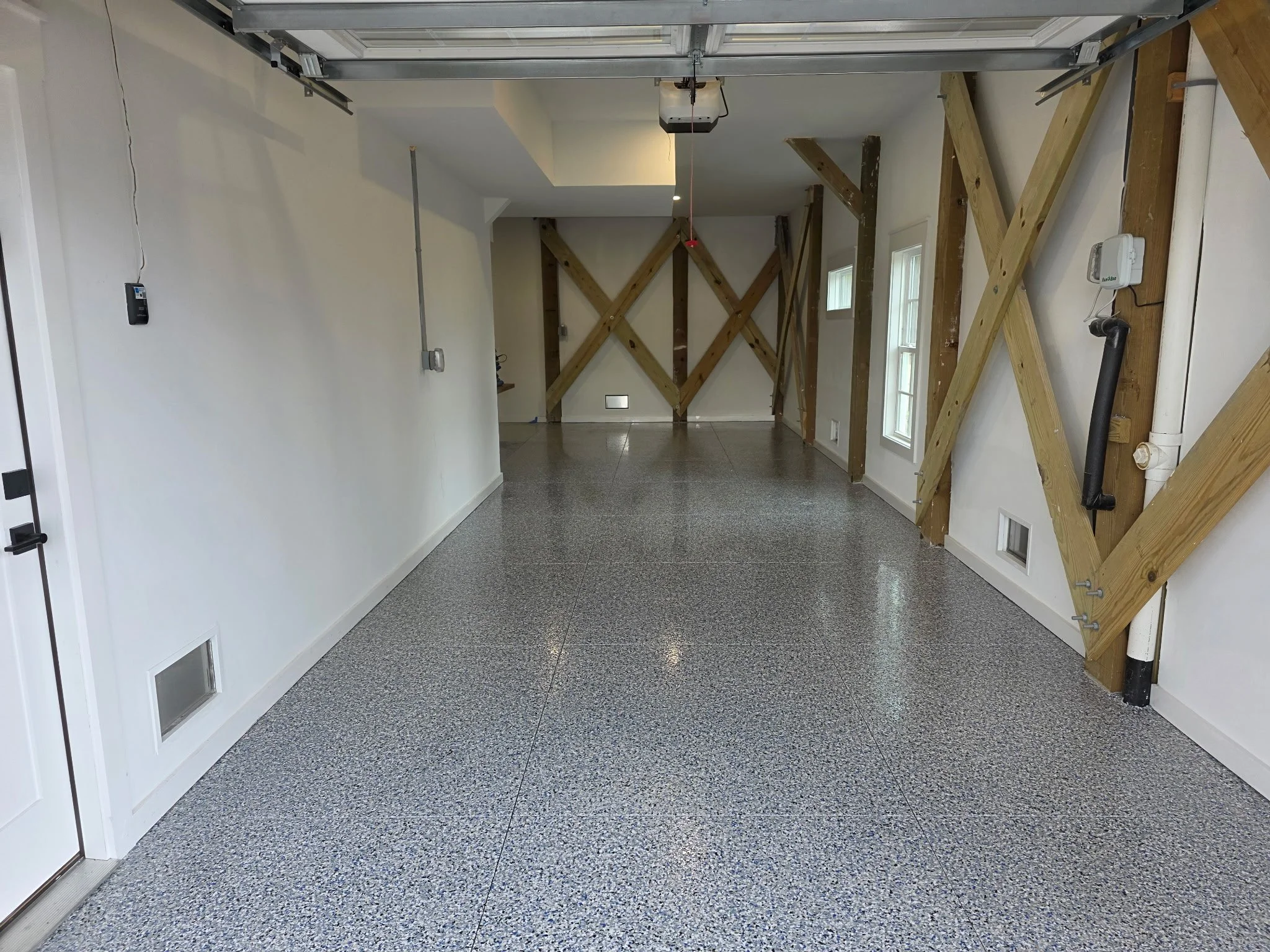 An indoor garage with white walls, large windows, and a speckled gray epoxy floor. The garage door is open, revealing wooden support beams and a garage door opener mechanism. There are small wall-mounted utility devices and electrical outlets visible