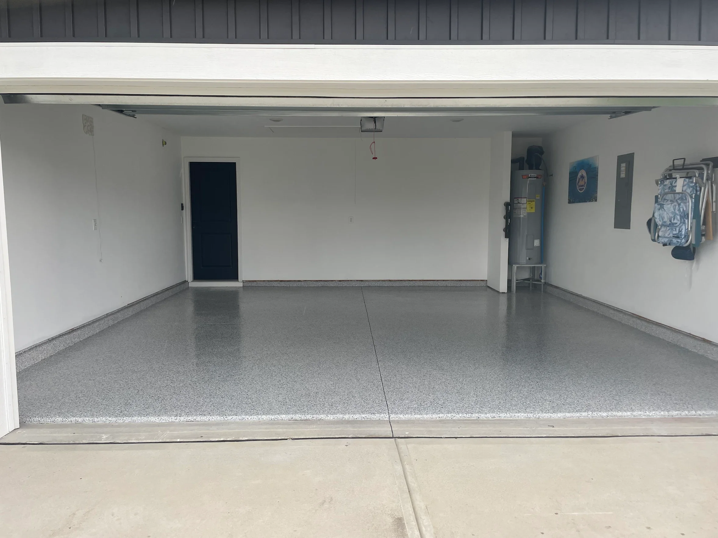 Open garage door revealing a clean, empty garage with white walls and terrazzo flooring. There is a water heater and power panel on the right side and a small painting on the wall. A backpack hangs on the wall, and a ladder leans against the right wall.