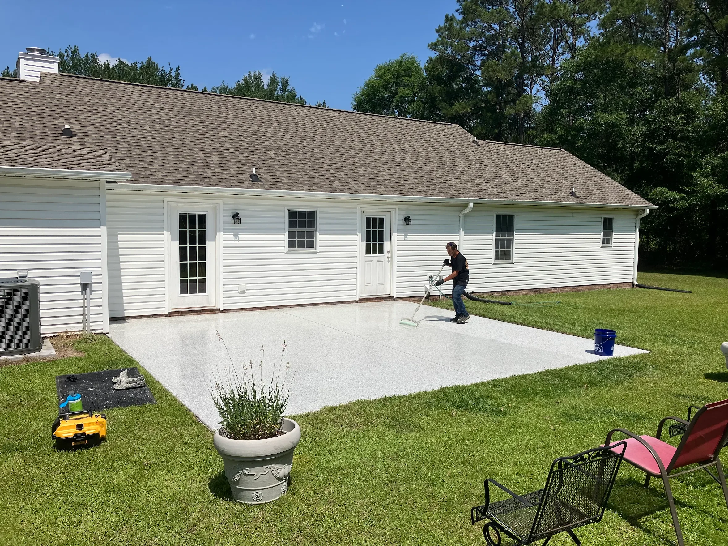 A person cleaning a patio with a power washer on a sunny day. The house has white siding, several windows, and a brown shingle roof. There are chairs and plants in the yard.
