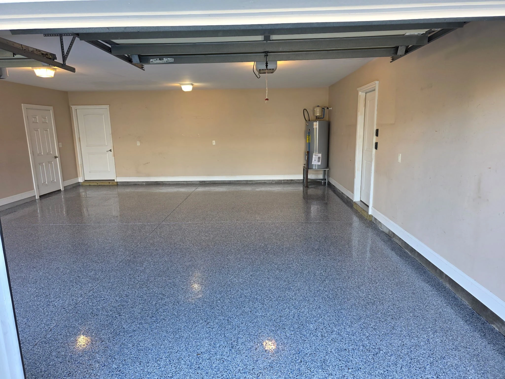 Empty garage with beige walls, white doors, and a gray speckled epoxy floor. There is a water heater in the corner and an open garage door.