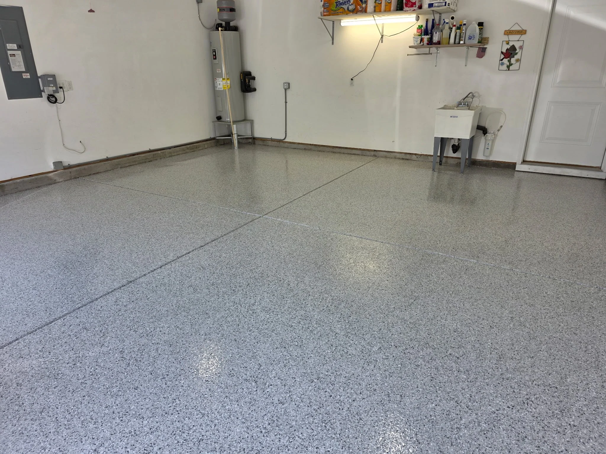 Clean garage with speckled gray epoxy floor, electrical panel, water heater, shelves with household items, laundry sink, and a closed white door.