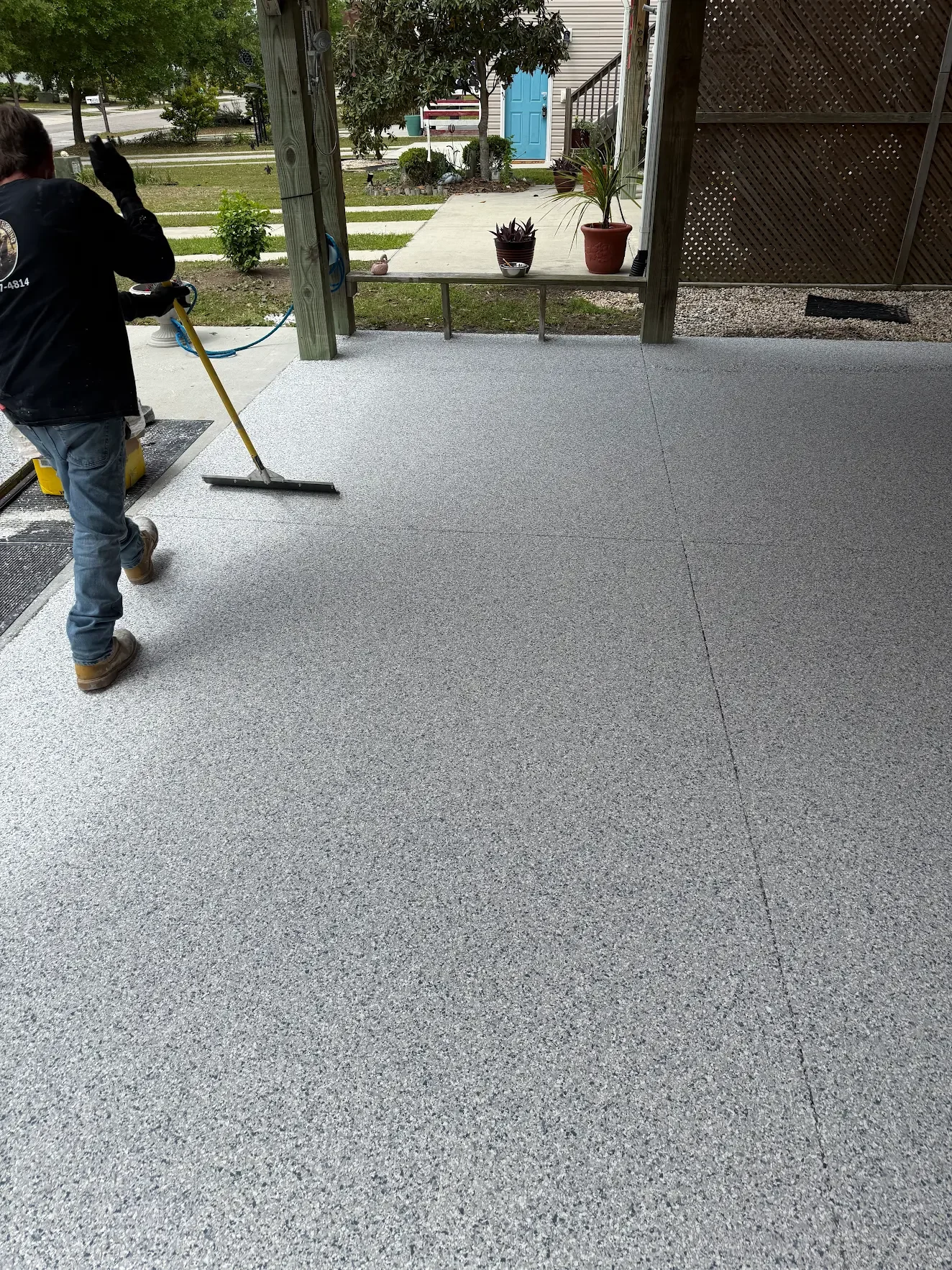 A person in work clothes is sweeping a textured concrete patio. The background shows a grassy yard with potted plants, a walkway, and a house with a bright blue door.