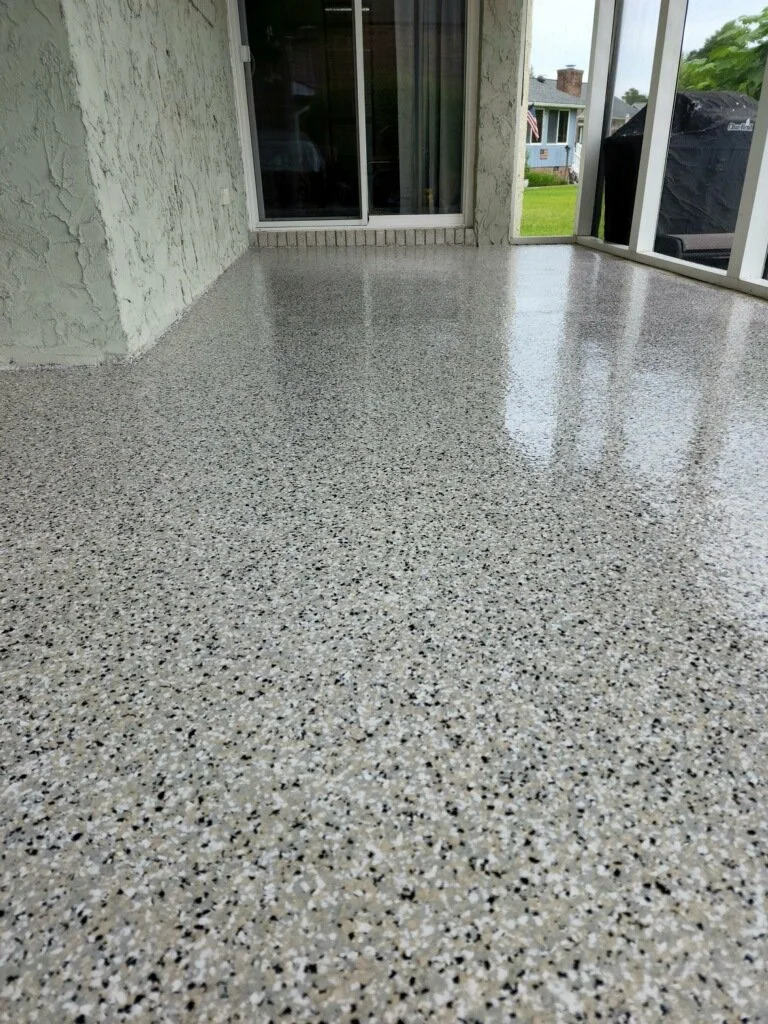 Porcelain or epoxy coated outdoor patio with speckled beige, black, and white floor surface, enclosed by screened walls with a sliding glass door leading to the interior, and visible in the background are houses, grass, and a grill.