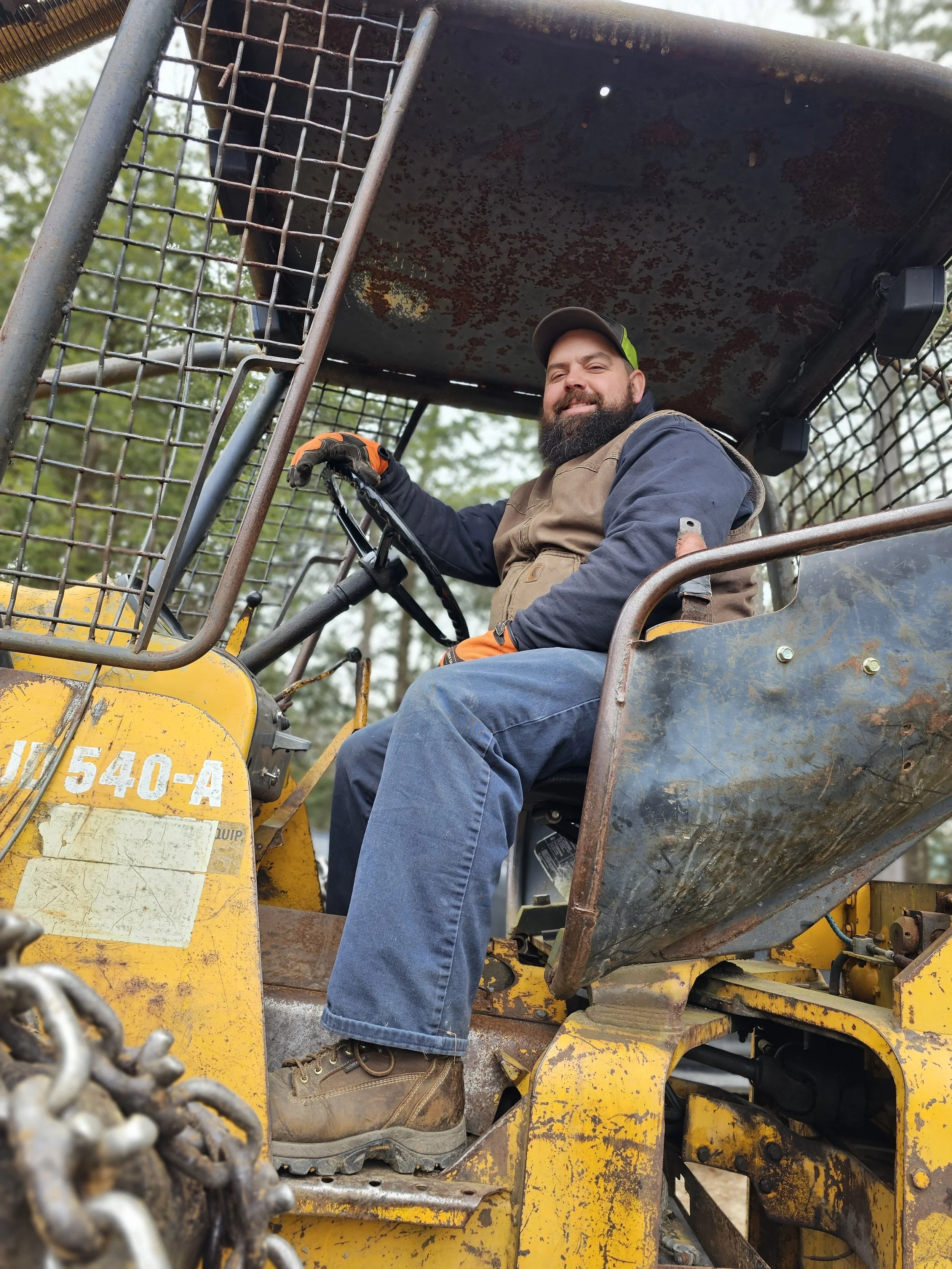 A man with a beard and a cap smiling while sitting in a yellow industrial tractor, with a rusted metal roof and grid-like metal barriers in front.