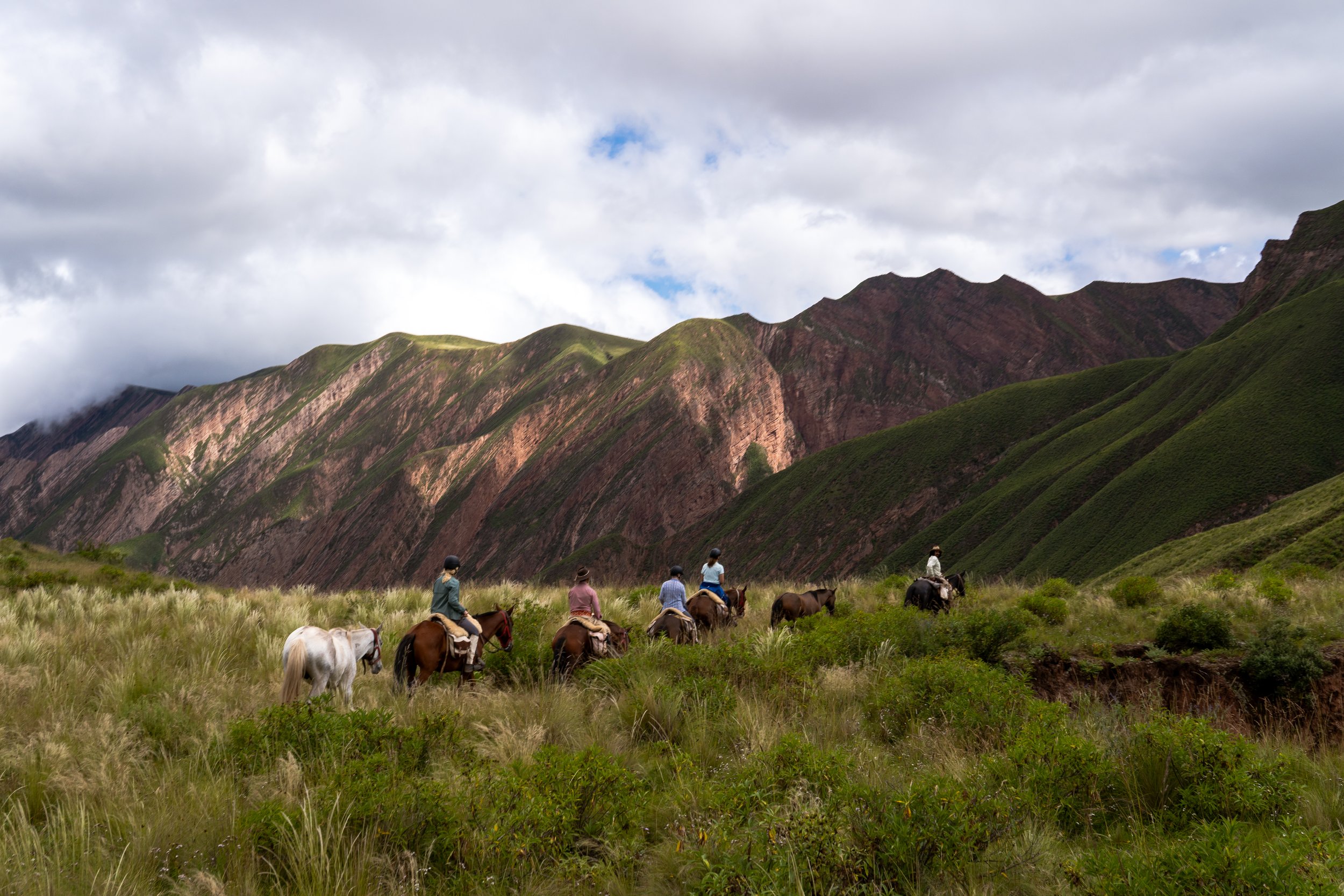 A group of people riding horses through green grassland with mountain ranges in the background under a cloudy sky.
