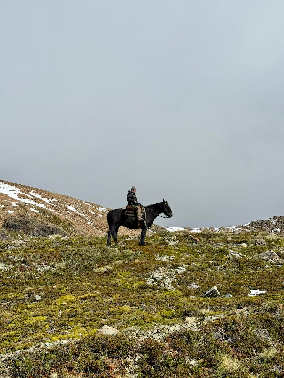 Person riding a black horse on a grassy, rocky hillside with patches of snow, under a cloudy sky.