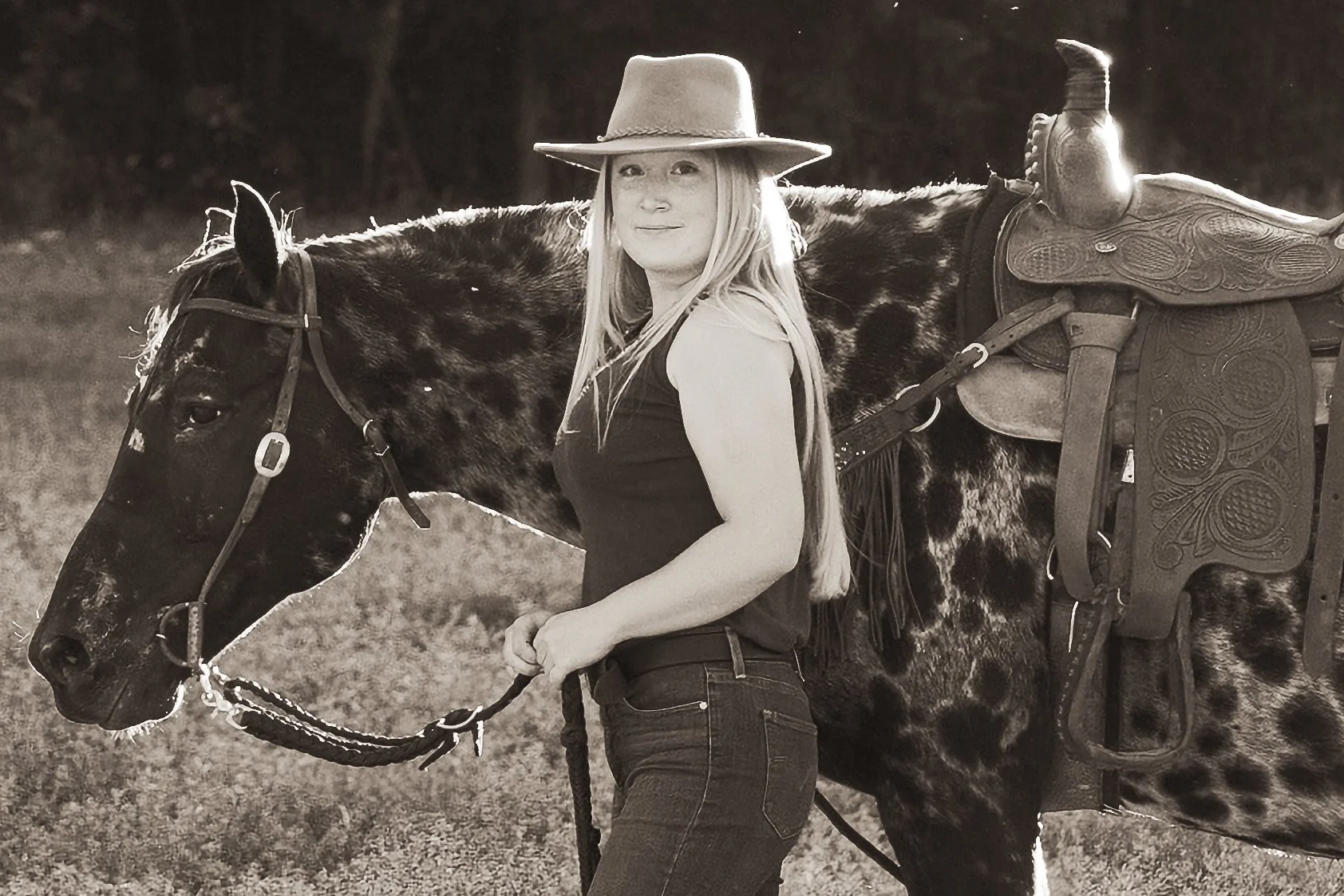 A woman with long hair, wearing a cowboy hat, stands next to a horse in an outdoor setting, holding the horse's lead rope.