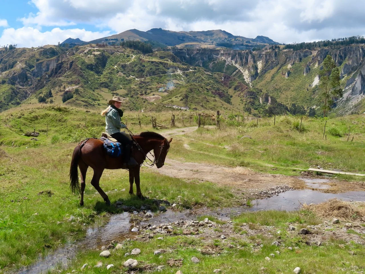 A woman riding a horse along a dirt path in a green mountainous landscape with cliffs and a cloudy sky in the background.