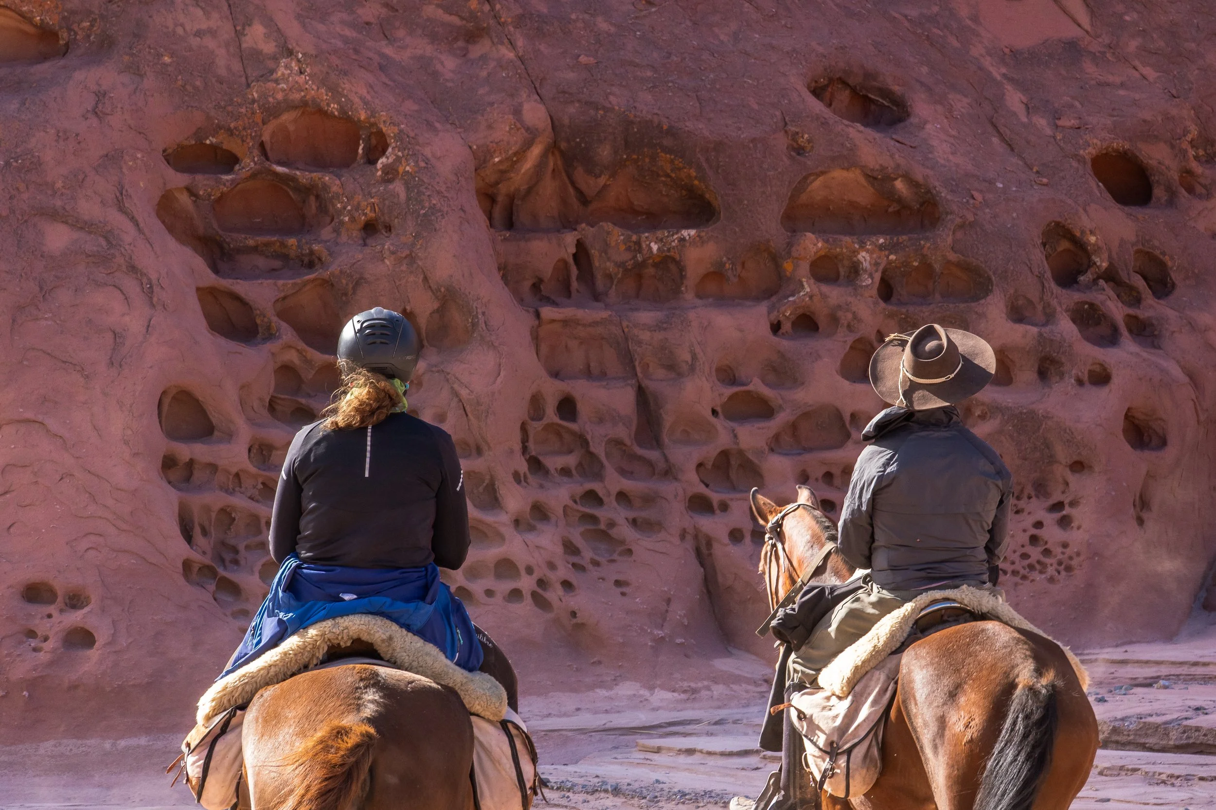 Two people on horseback are looking at a large, red rock formation with numerous holes on its surface.