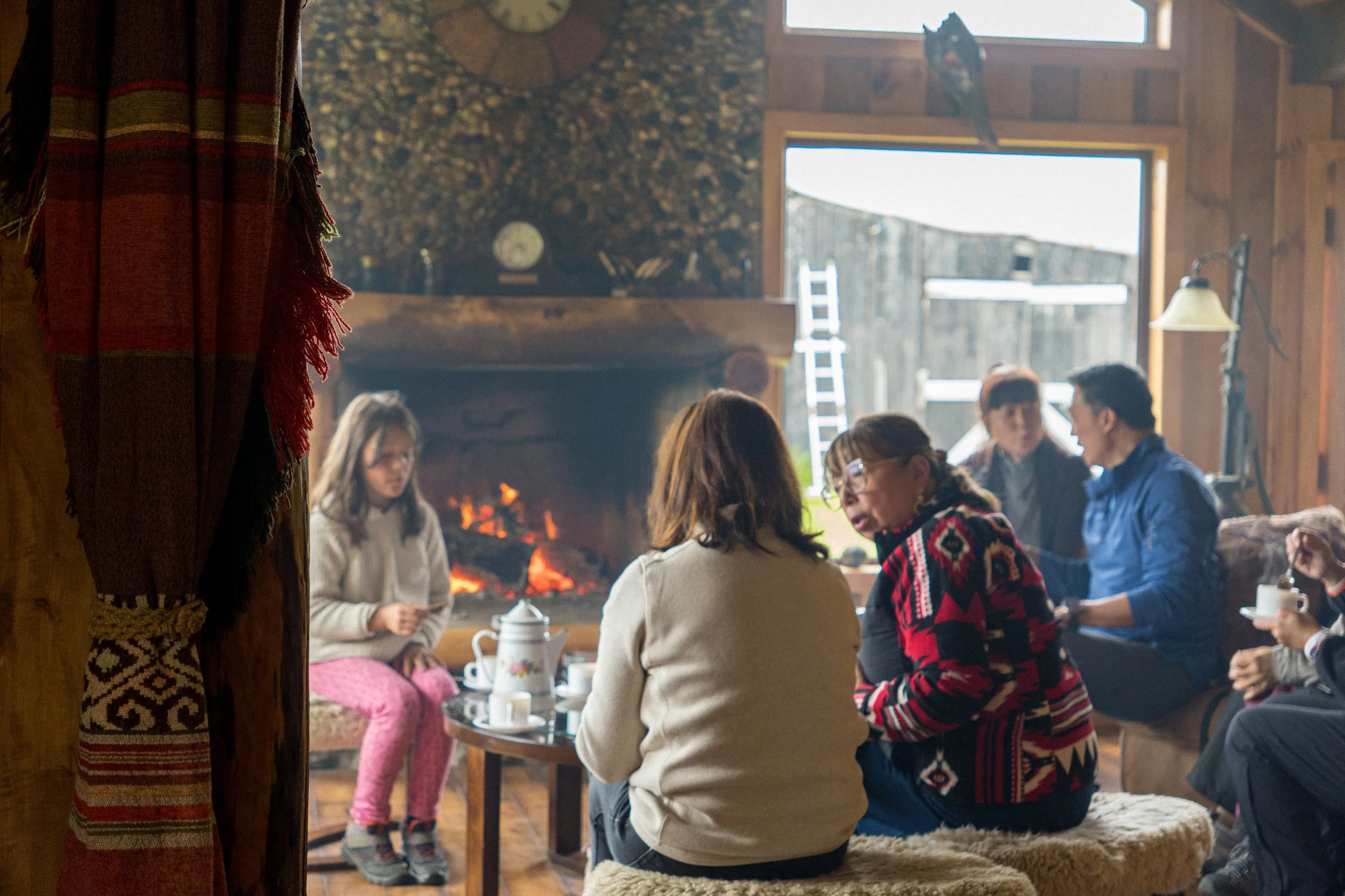 People sitting around a table having conversation inside a rustic cabin with a brick fireplace and wood walls. The scene appears warm and cozy.