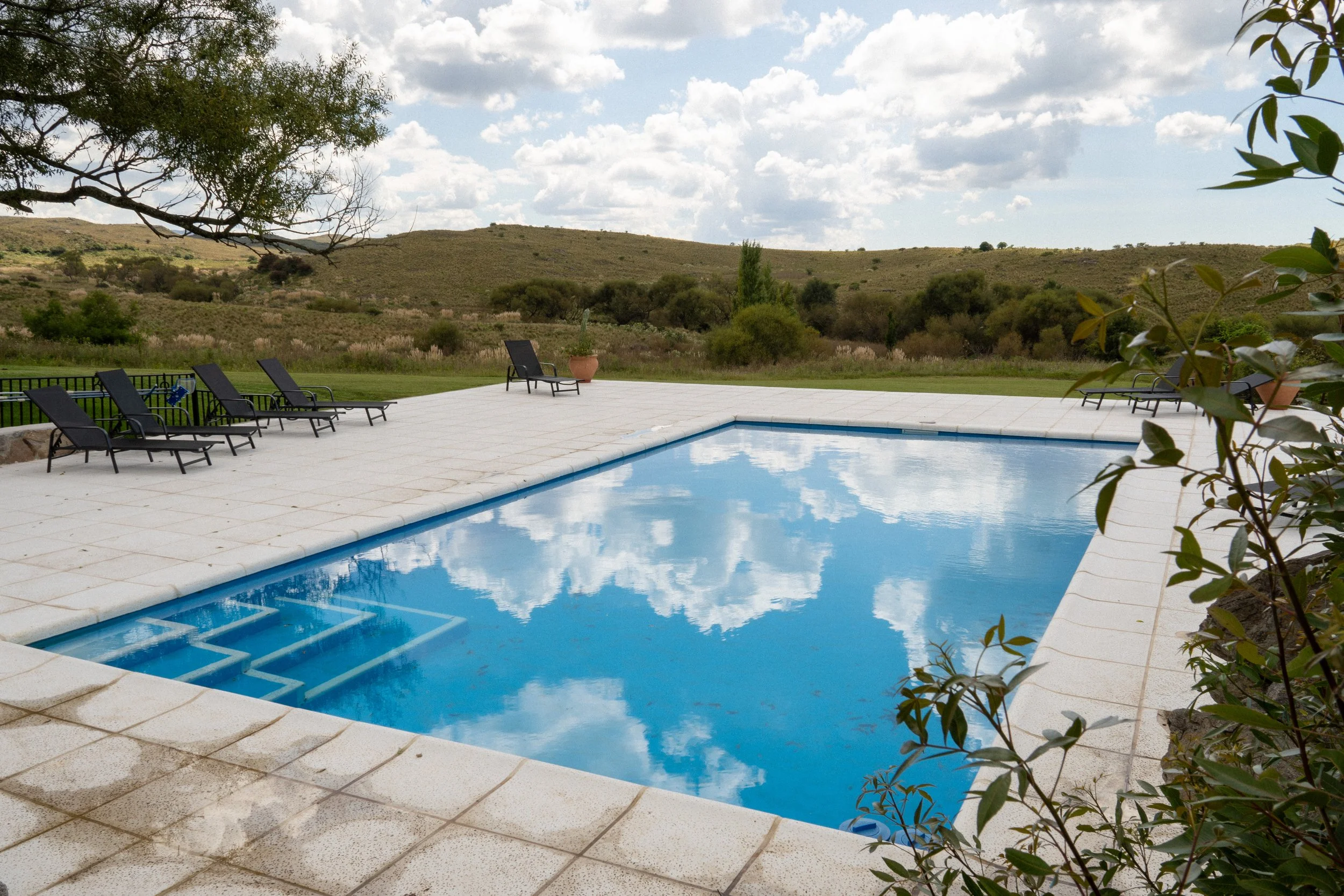 An outdoor swimming pool with lounge chairs around it, overlooking a landscape of rolling hills and partly cloudy sky.