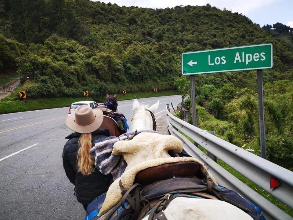 A person walking beside a horse along a mountain road near a sign that reads 'Los Alpes'. The rider wears a wide-brimmed hat and has long blond hair. Green hills surround the road.