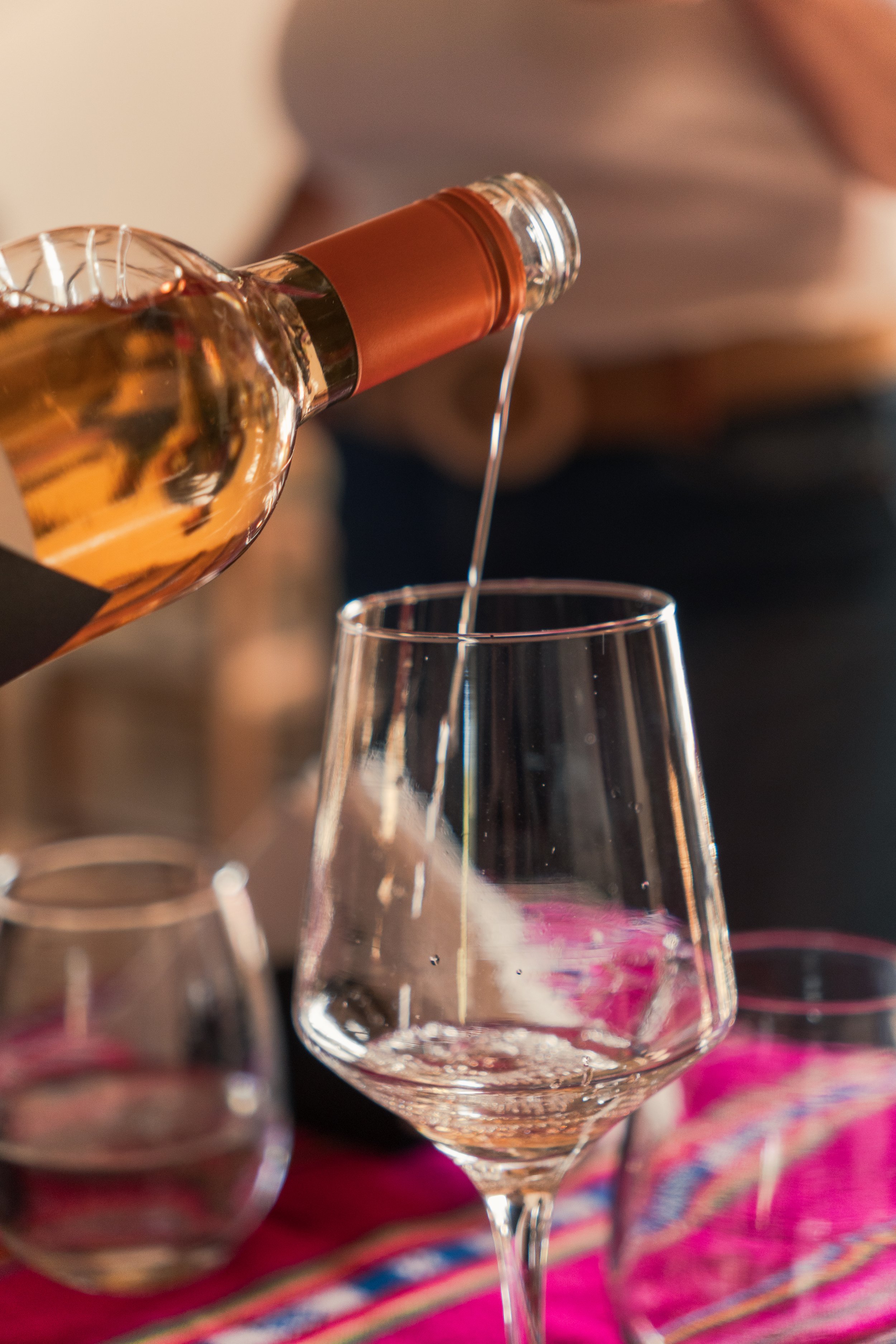 A person pouring a glass of white wine in a glass with a pink and purple patterned cloth underneath.