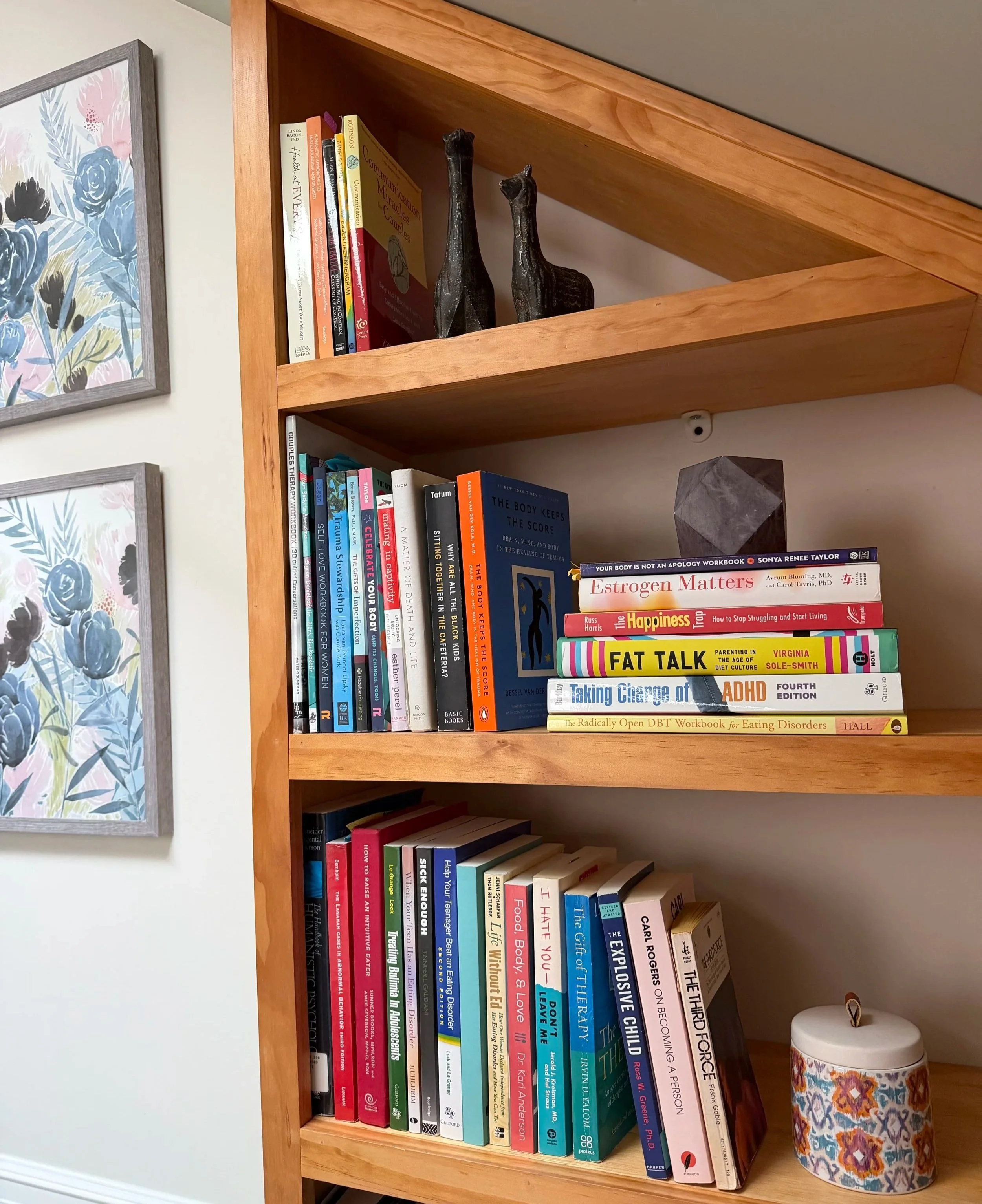 A wooden bookshelf containing a variety of therapy books, decorative giraffe sculptures, and a floral-patterned ceramic container, with framed floral artwork on the wall beside it.