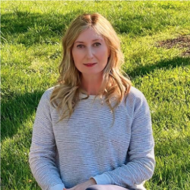 A woman with long blonde hair wearing a light gray sweater sitting outdoors on green grass.