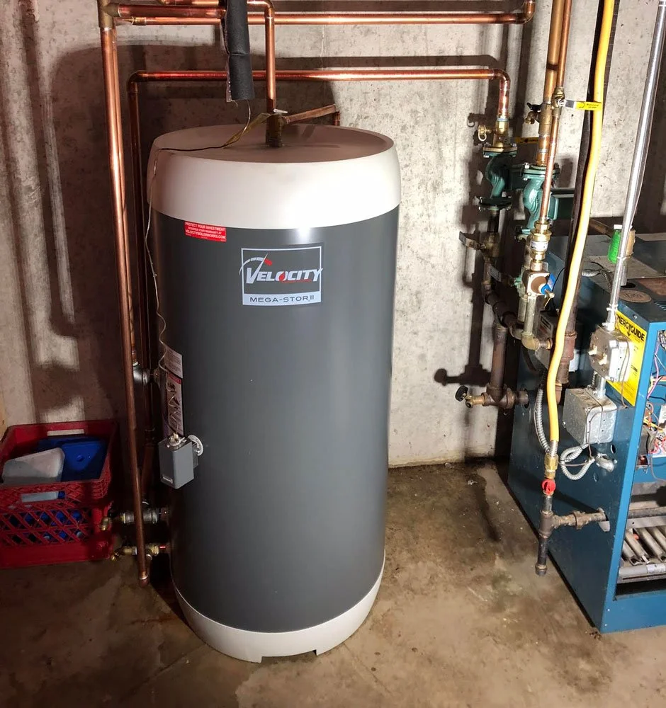 A hot water heater tank labeled VLT velocity Mega-Stor II, surrounded by copper pipes and connected to a plumbing and heating system in a basement or utility room.