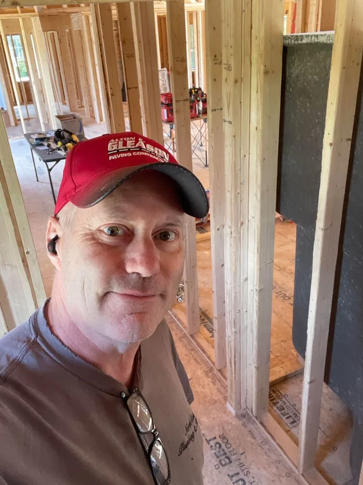 A man taking a selfie inside a house under construction, with exposed wooden framing and partially finished rooms.