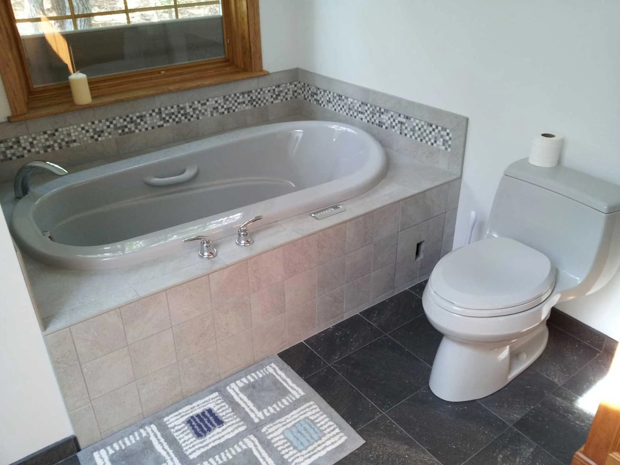Bathroom with a white bathtub under a window, a toilet with a roll of toilet paper, a gray patterned rug, and a tiled wall with a decorative mosaic border.