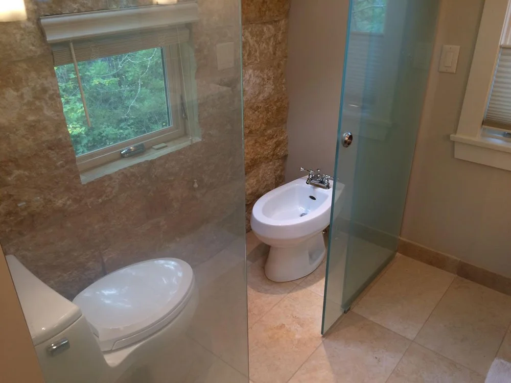 Bathroom with a small window, a toilet, as well as a bidet, separated by a glass panel, with tiled walls and floors.