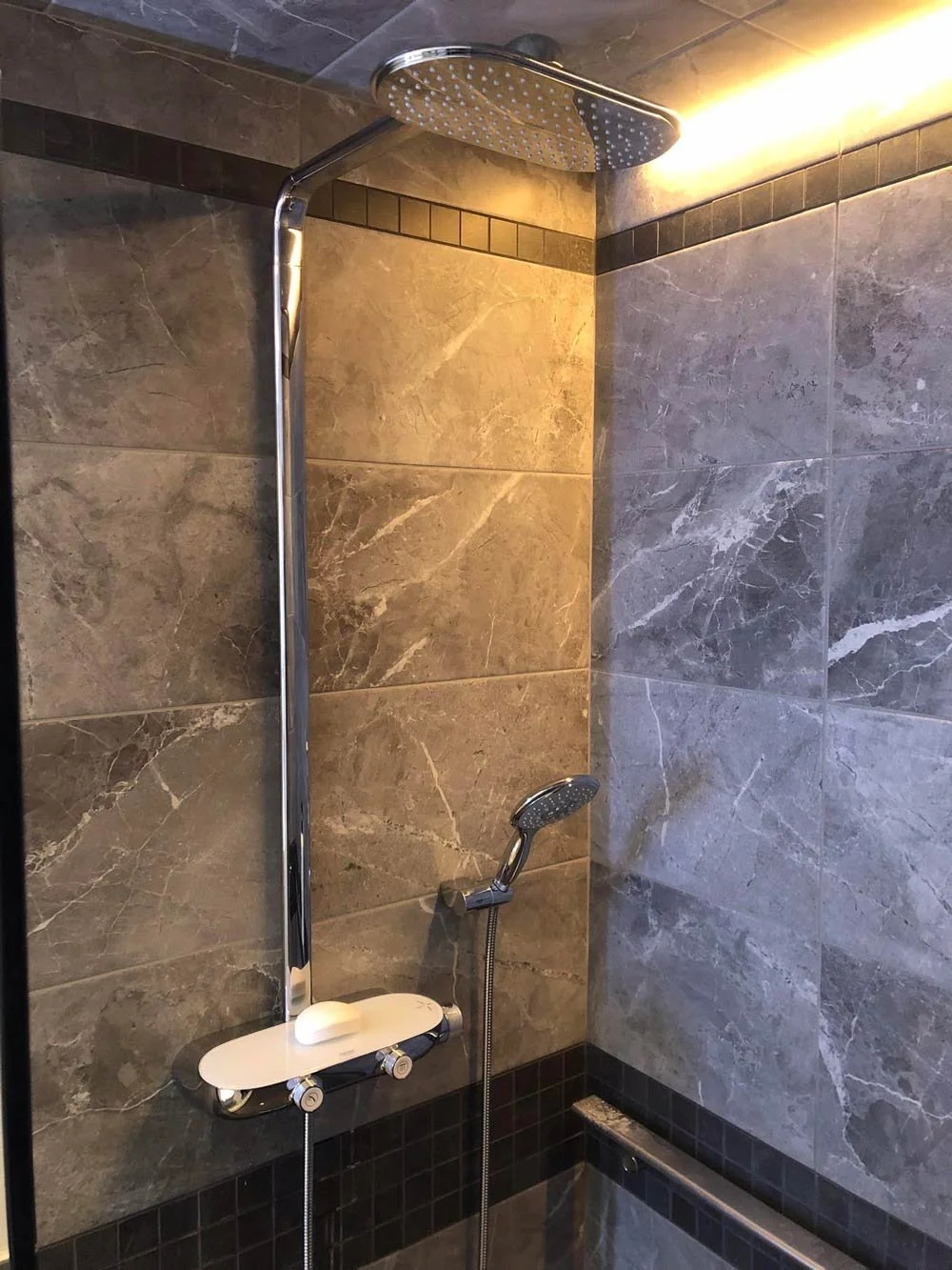 Shower area with a large rain shower head and a smaller handheld shower head, both attached to a chrome pipe. The wall is covered with large gray stone tiles, and there are small black tiles at the bottom. The shower controls are mounted on a white panel with two knobs.