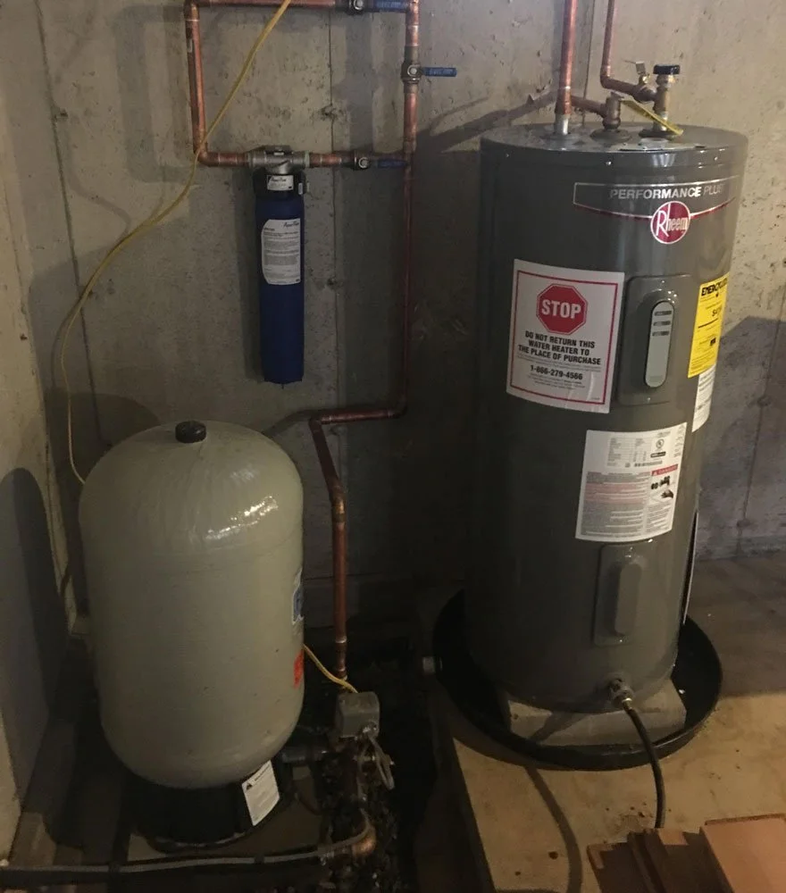 Water heater and water softener in a basement utility room, with pipes and a filter mounted on the wall.